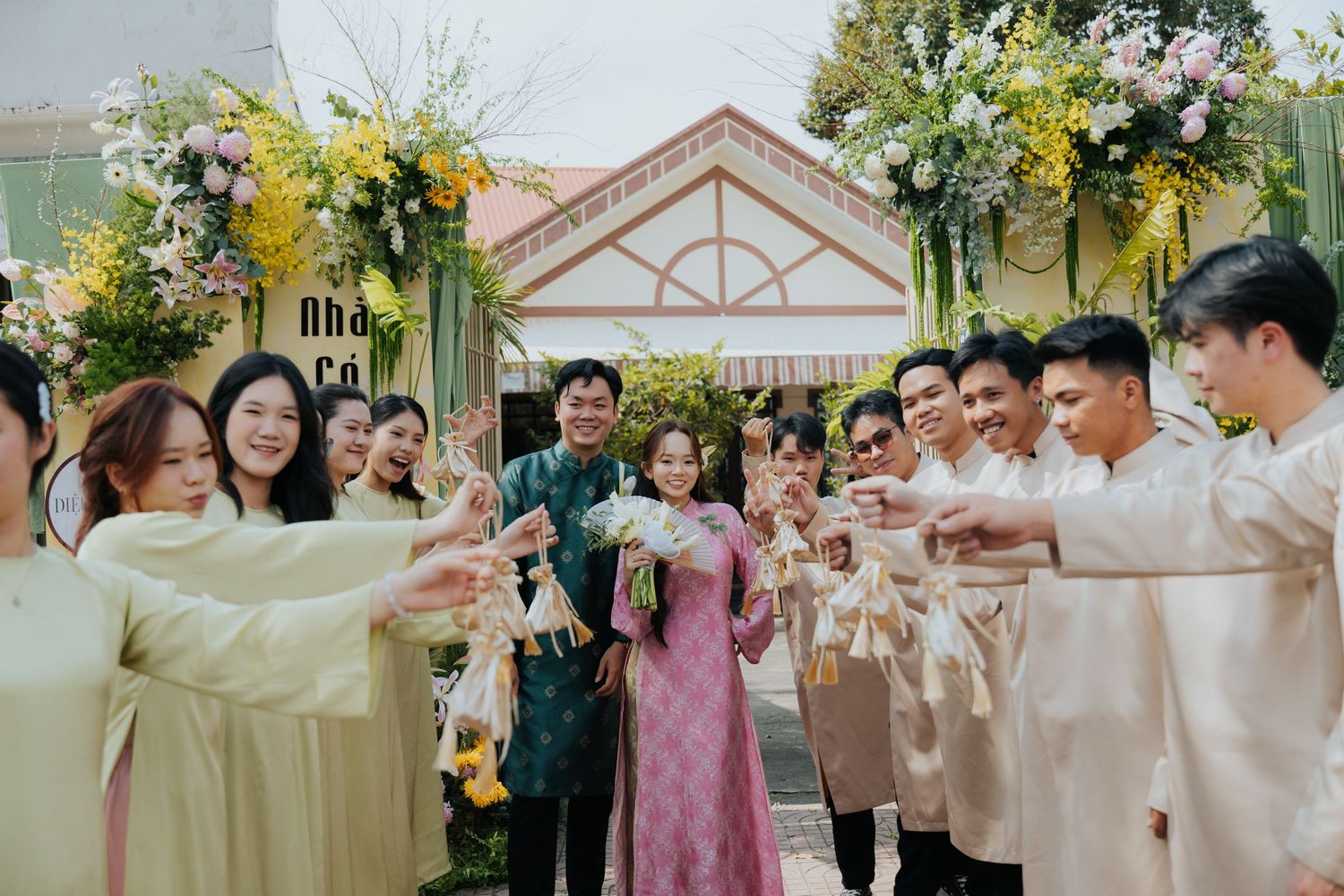 Vietnamese wedding ceremony with couple in traditional ao dai attire, surrounded by guests forming a celebratory arch outdoors.