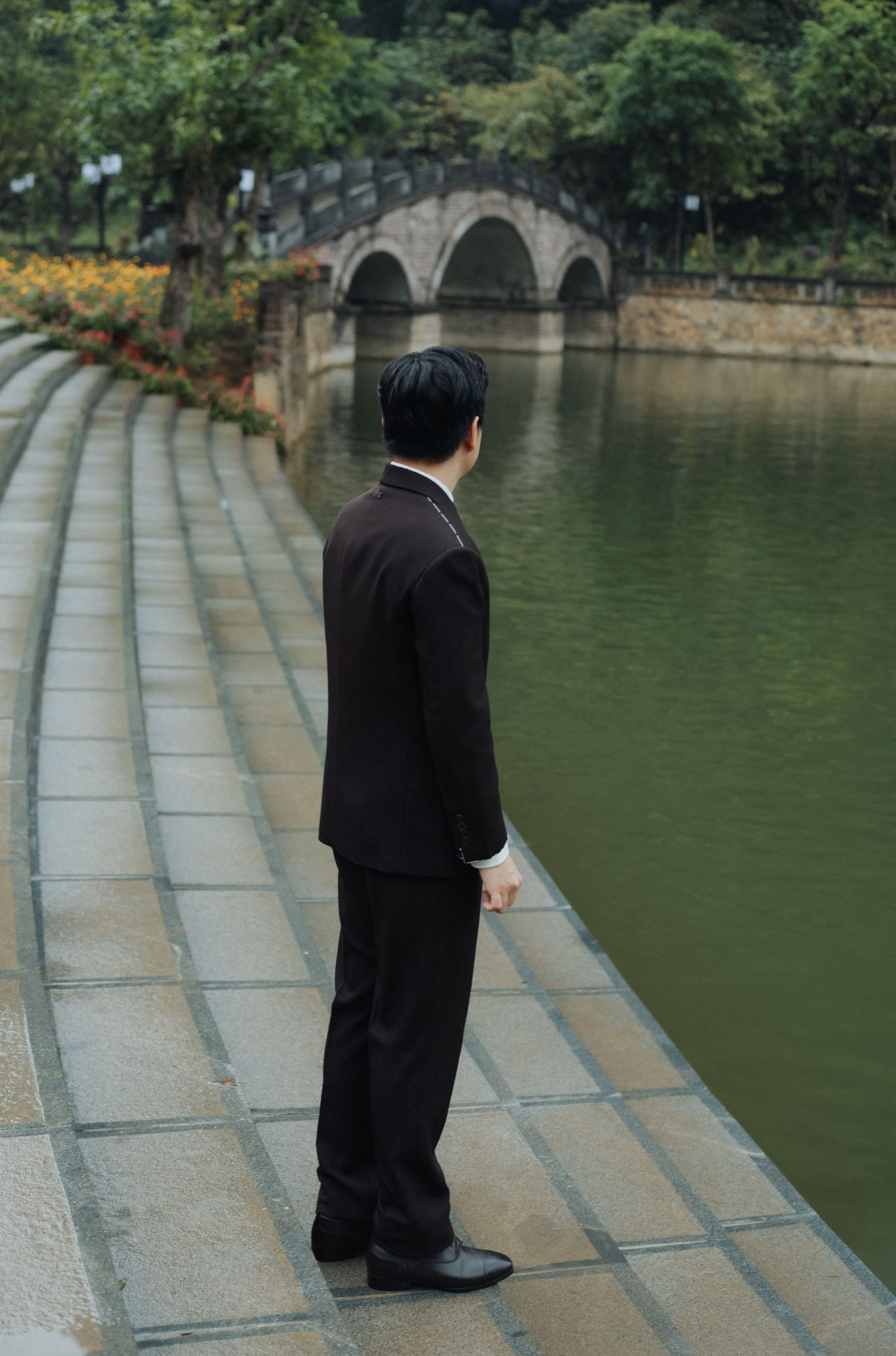 A man in a dark suit stands on stone steps beside a tranquil green pond with an arched bridge in the background.