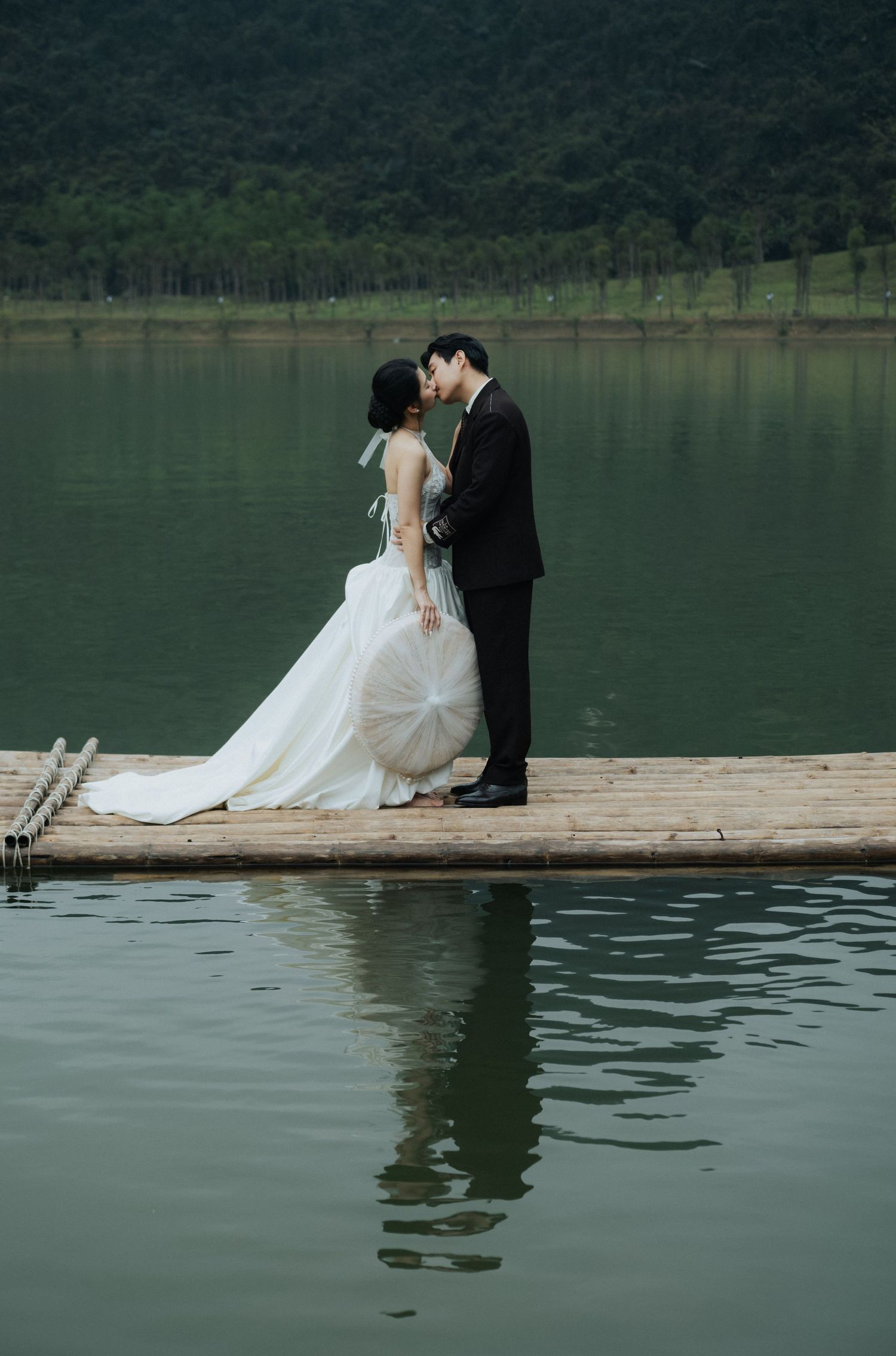 A couple shares a romantic kiss on a wooden dock over a serene lake, bride in white gown, groom in black suit.