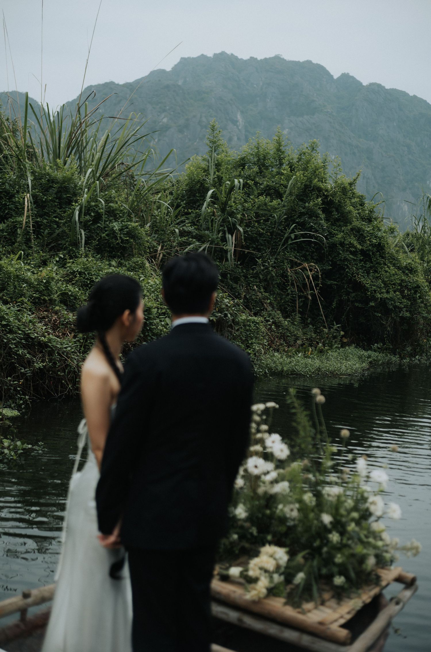 Couple in wedding attire standing on a bamboo boat with flowers, overlooking a misty river and lush green mountains.
