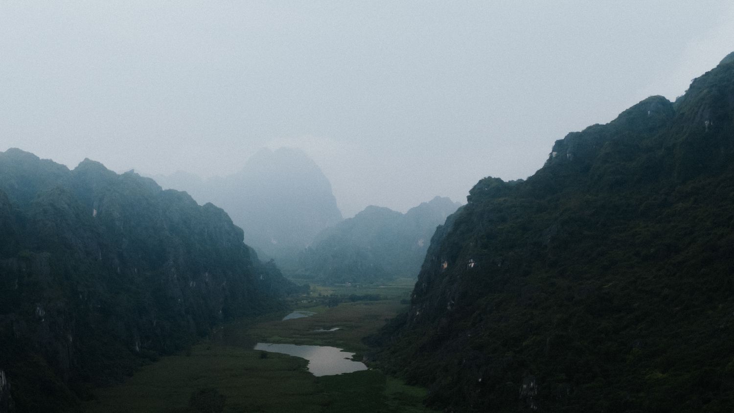 Misty karst mountains surround a lush green valley in Vietnam, with dramatic limestone peaks shrouded in fog.