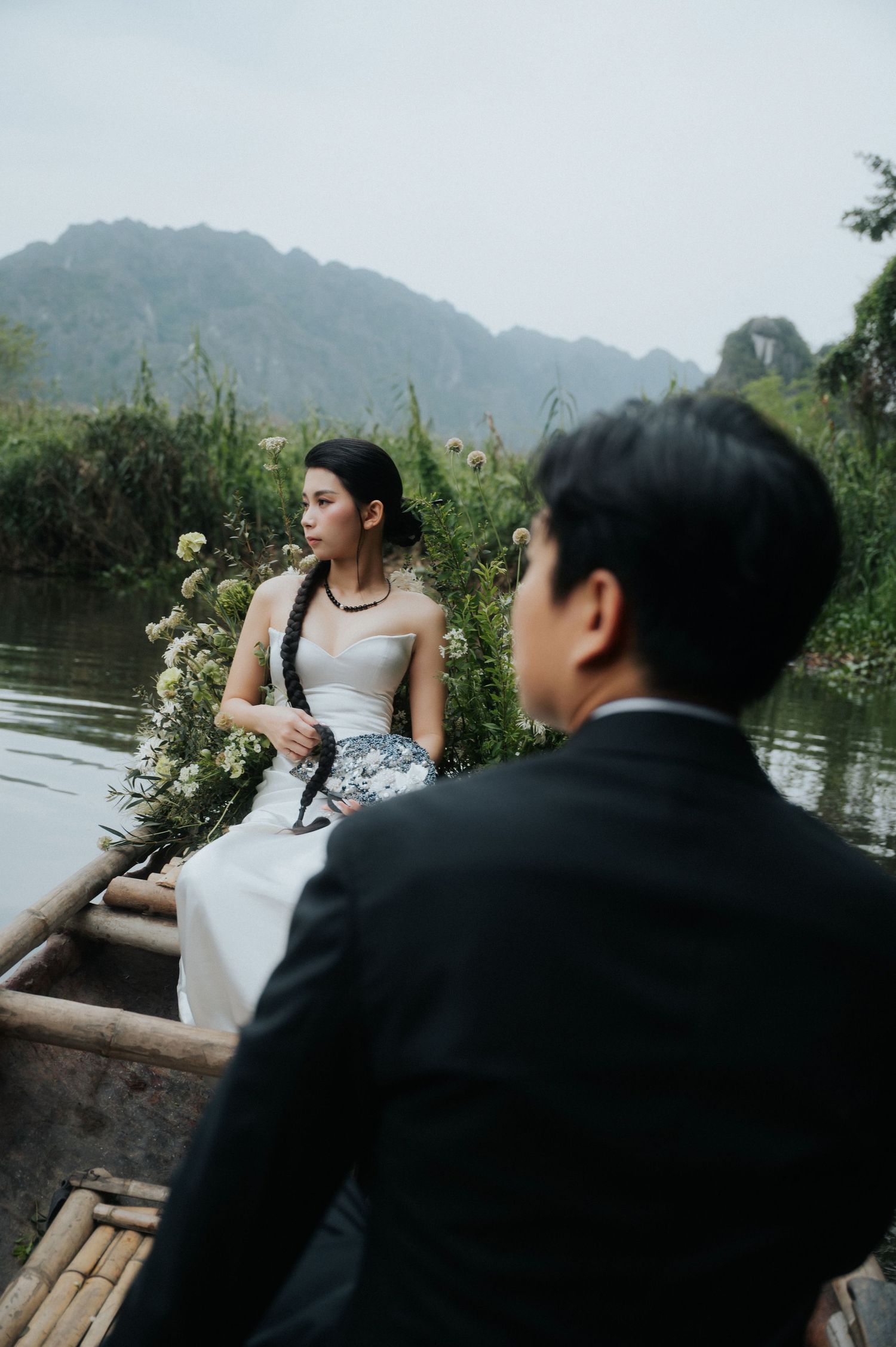 A bride in white dress holds flowers on a wooden boat with her groom in a suit, surrounded by lush greenery and misty mountains.