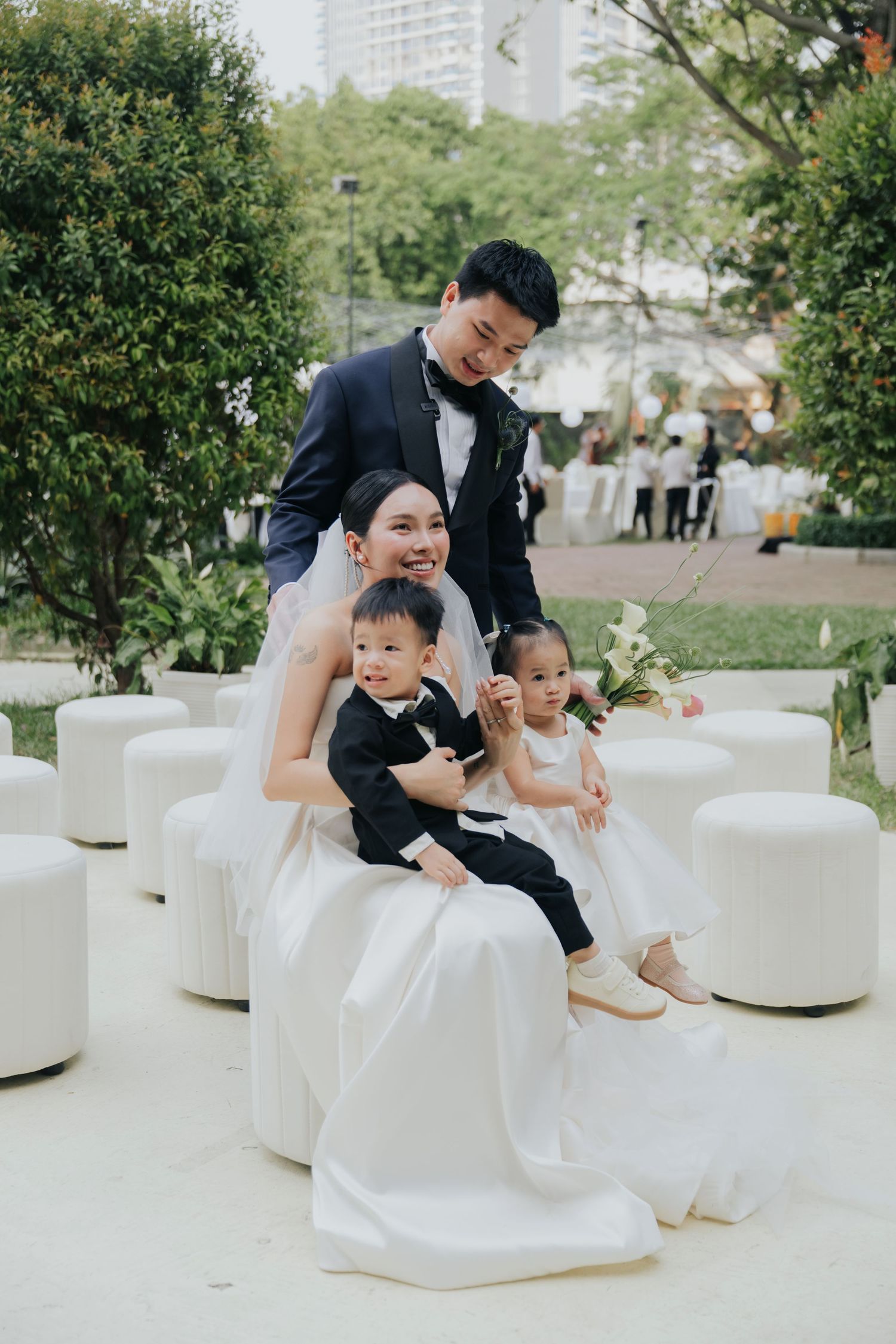 Bride and groom pose with two young children in formal attire at an outdoor garden wedding venue.