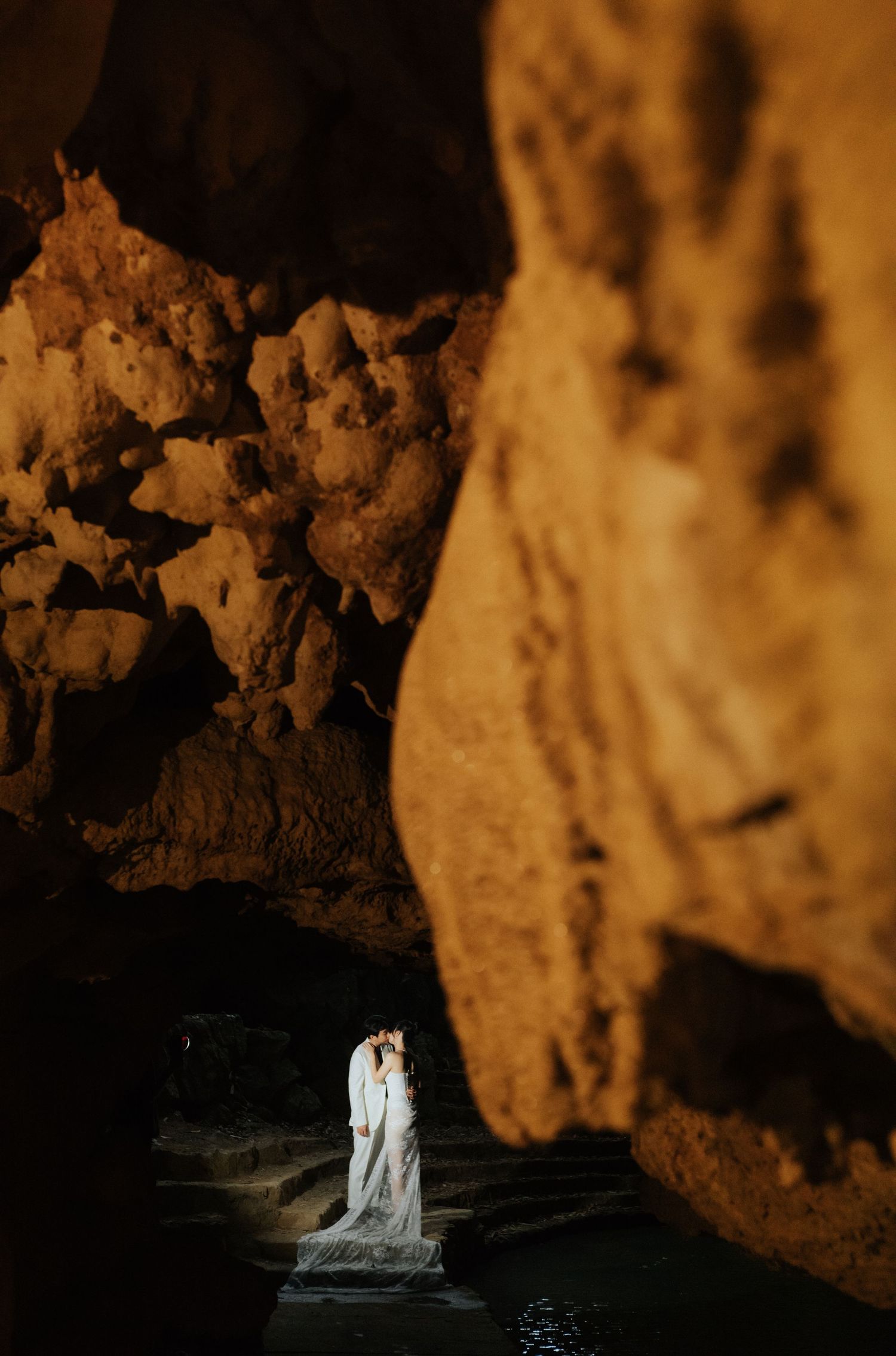 A couple shares a romantic kiss in a dramatic cave setting, illuminated by warm golden light against rugged rock walls.