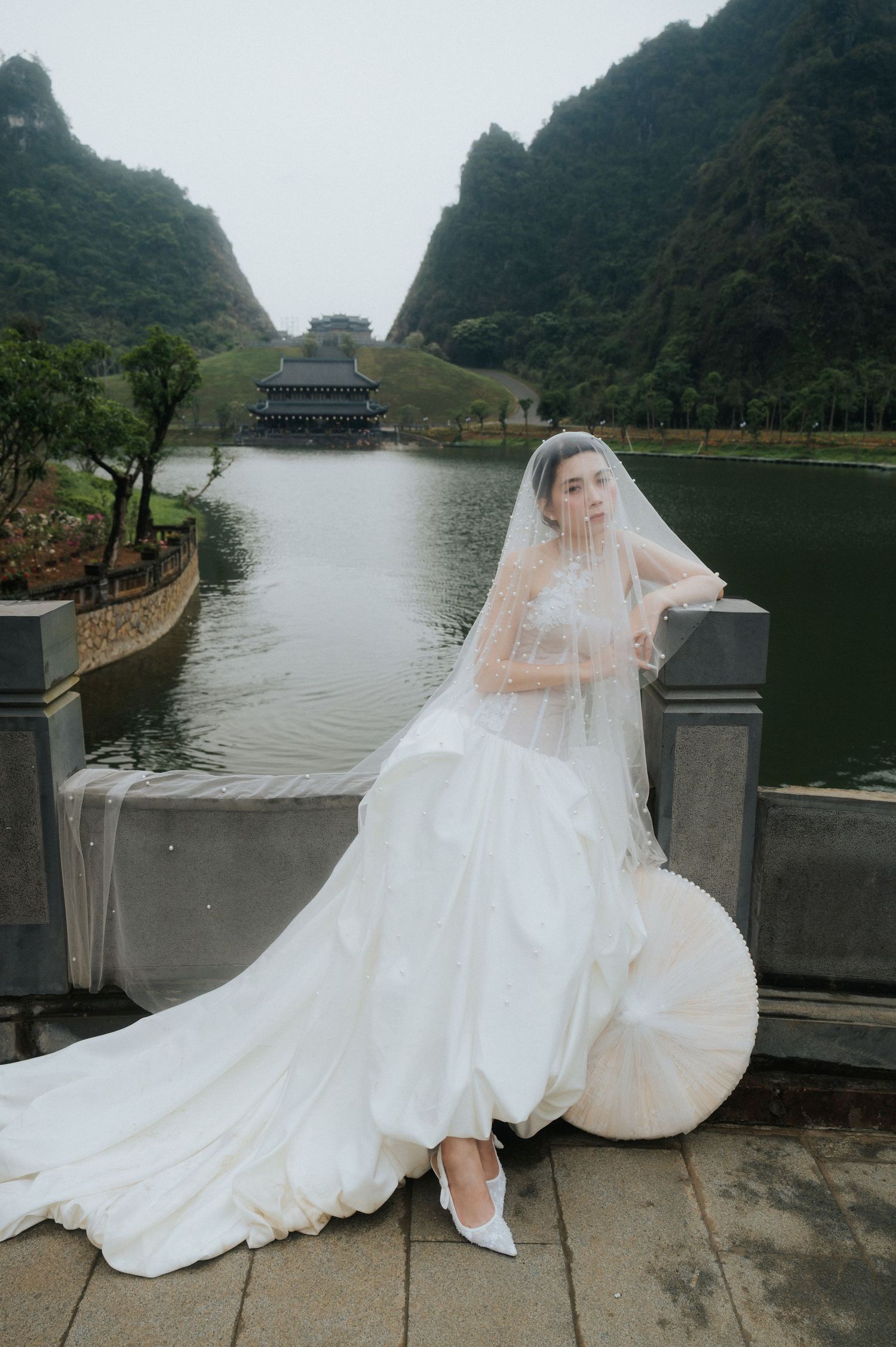Bride in white wedding gown and veil poses on stone bridge near tranquil lake with pagoda and green mountains backdrop.
