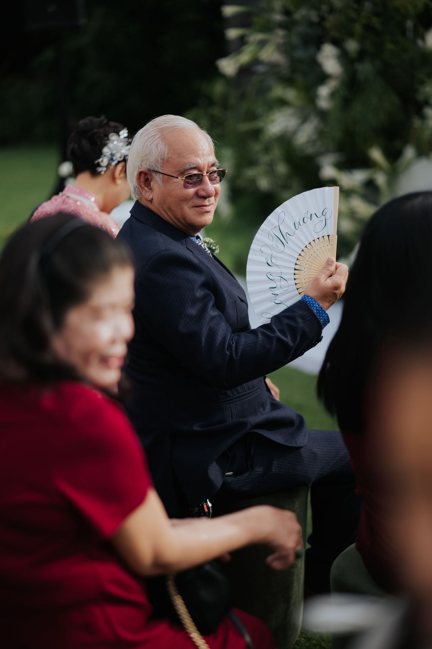 Elderly man in suit holding a decorative hand fan at an outdoor wedding ceremony.