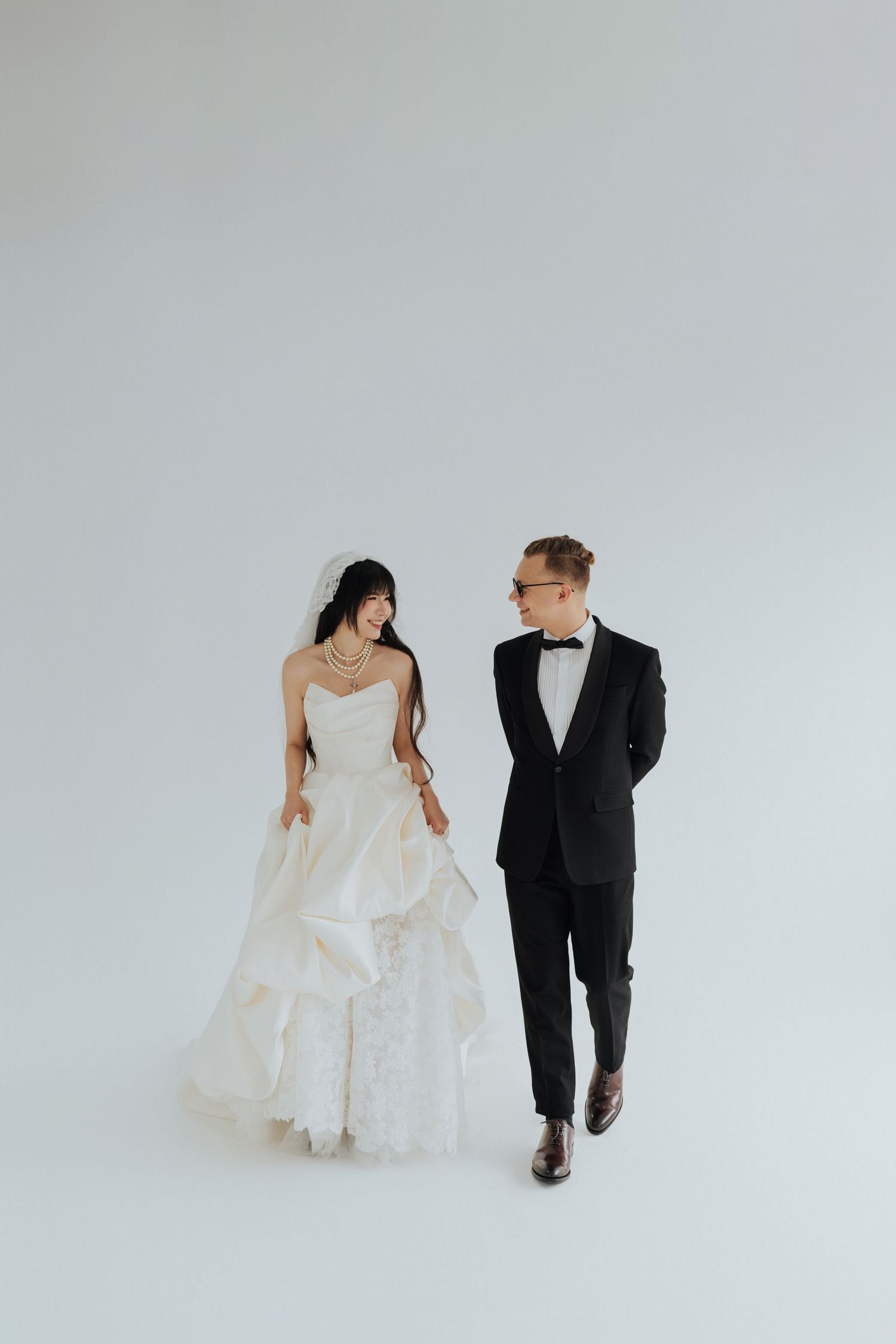 A bride in a white ballgown and groom in a black tuxedo walk together against a light gray studio background.