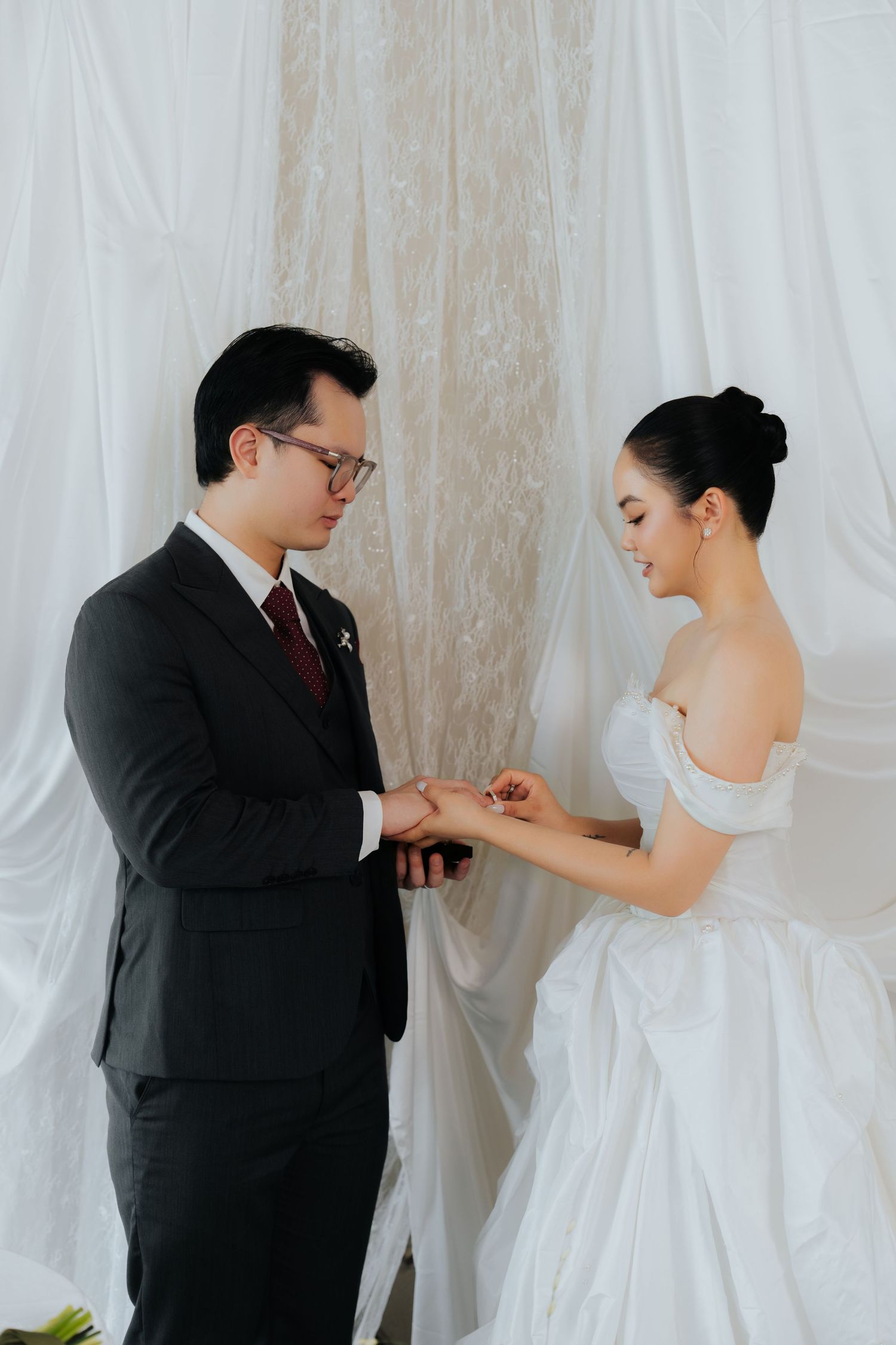 A bride in a white off-shoulder gown places a ring on the groom's finger during their wedding ceremony.
