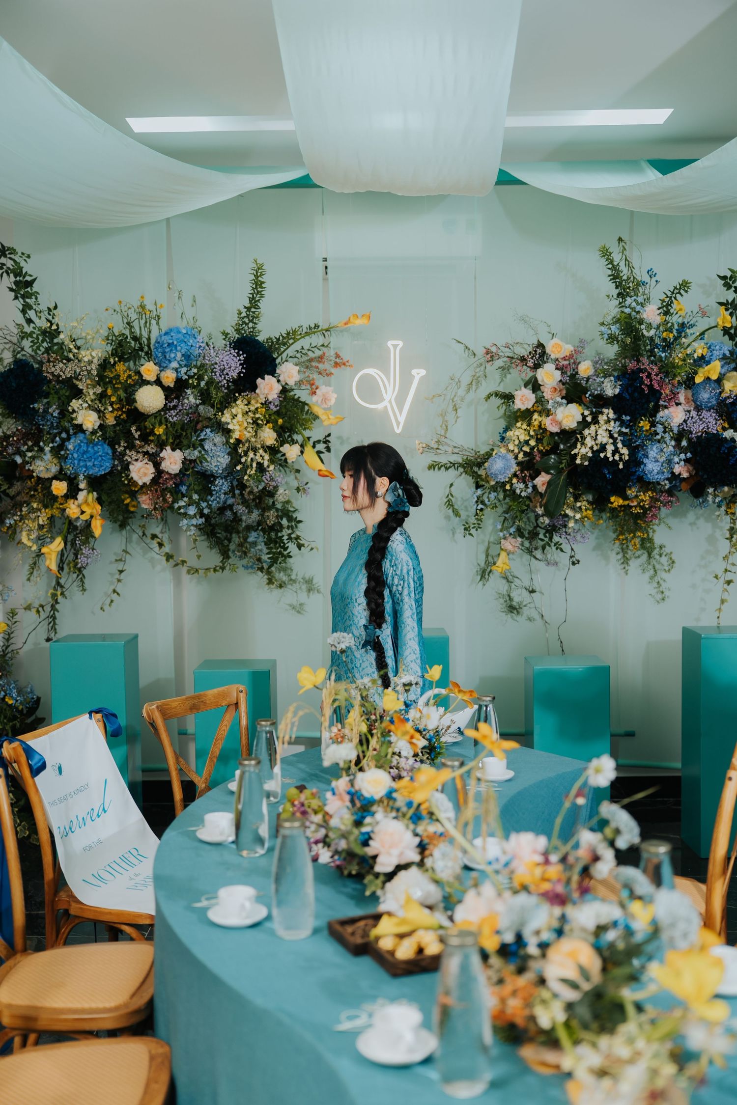 Elegant teal wedding reception with floral arrangements, a woman in blue dress, and a neon monogram backdrop.