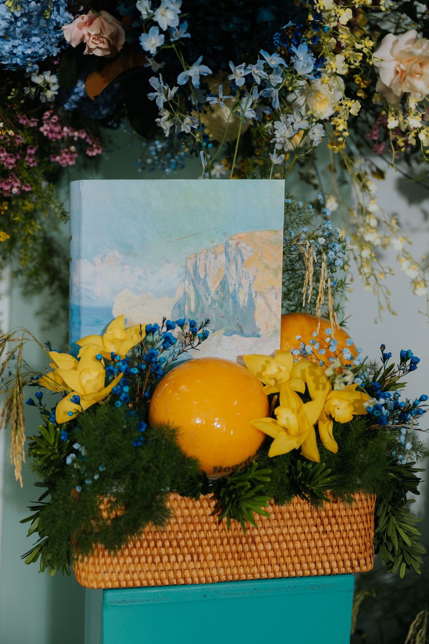 Decorative floral arrangement with yellow lilies, blue flowers, orange fruit, and a landscape painting in a wicker basket.