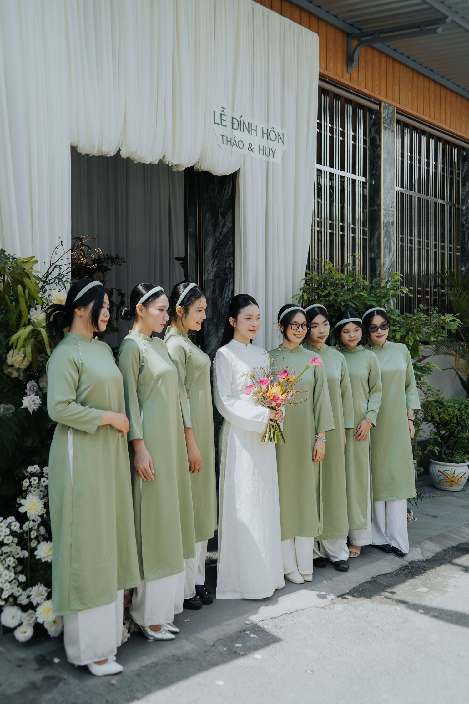 Vietnamese bride in white ao dai holds bouquet with bridesmaids in sage green ao dai at engagement ceremony.