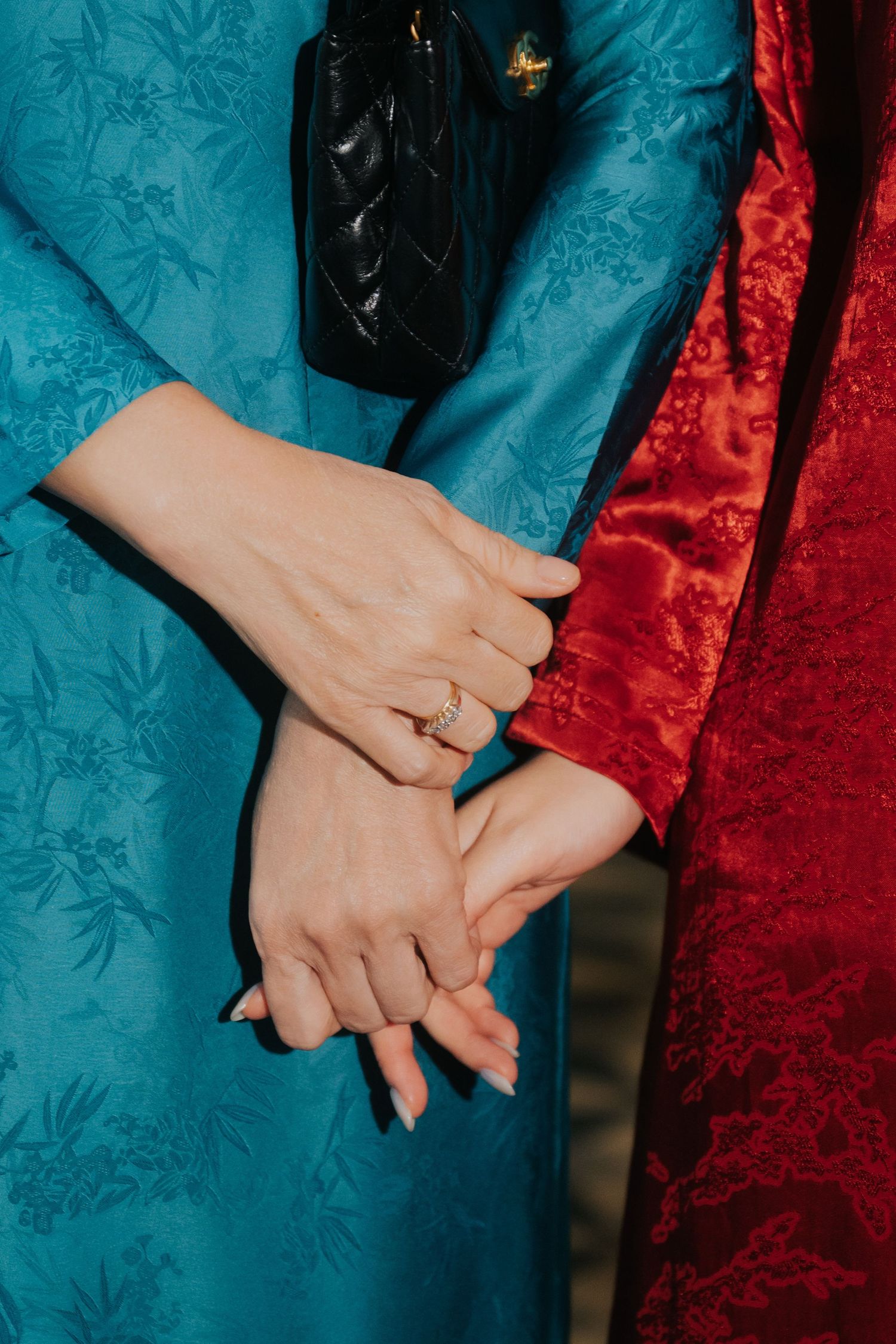 Two women in blue and red brocade dresses hold hands, one wearing a gold diamond ring, with a black quilted bag.