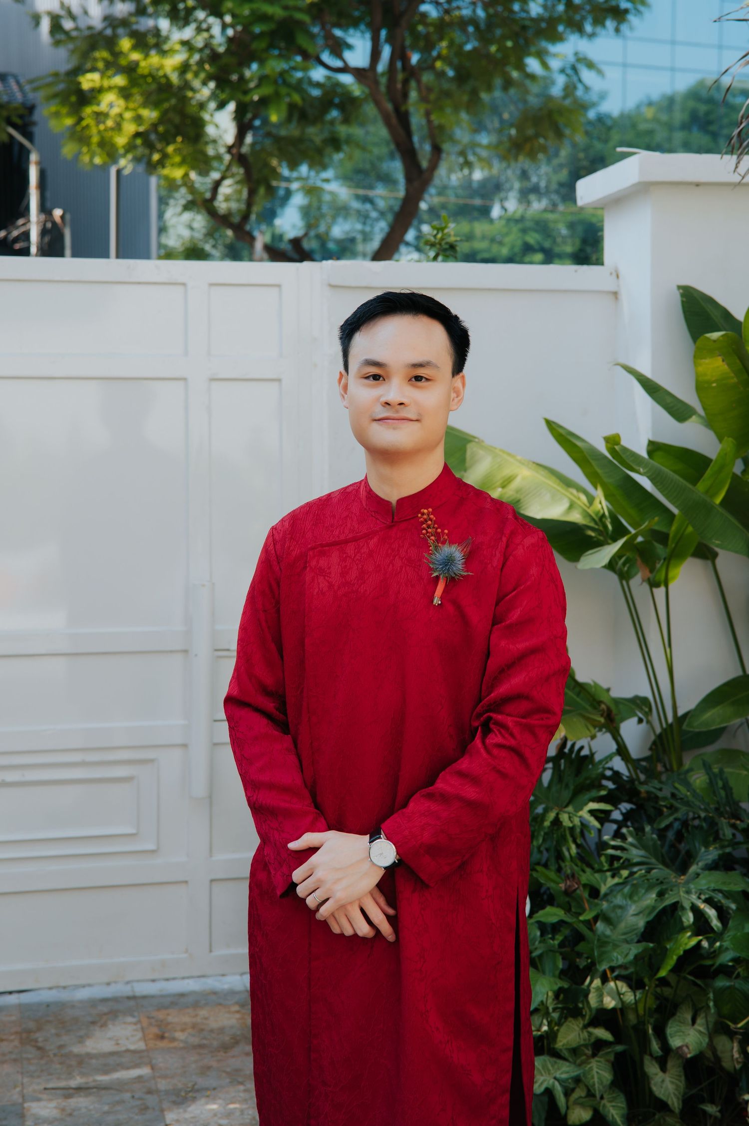 Young man wearing traditional red Vietnamese Ao Dai outfit, standing outdoors near white gate and tropical plants.