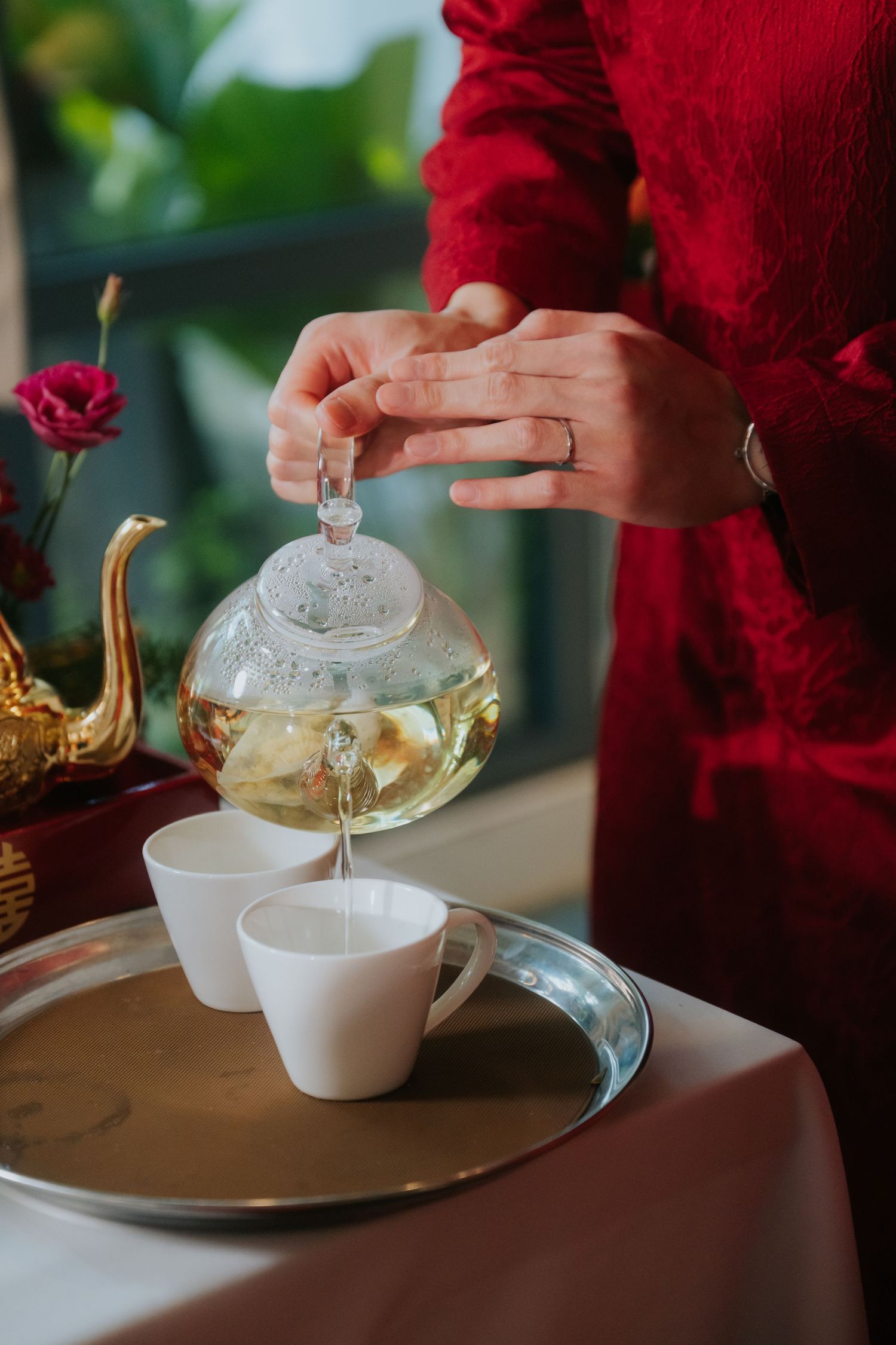 Woman in red dress pouring herbal tea from glass teapot into white cups on silver tray at elegant table.