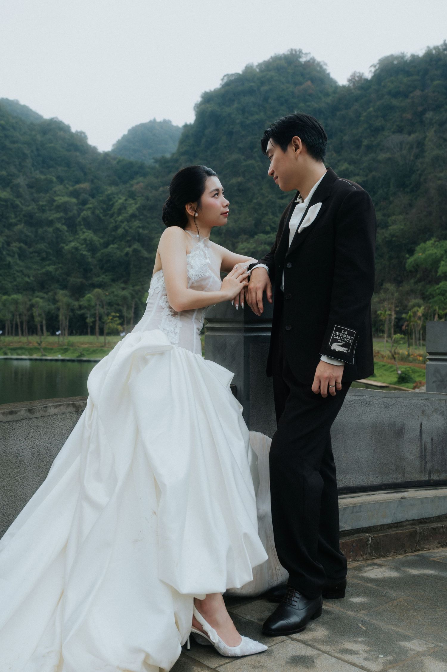 A bride in a white gown and groom in a black suit share a romantic moment outdoors with lush green mountains behind them.