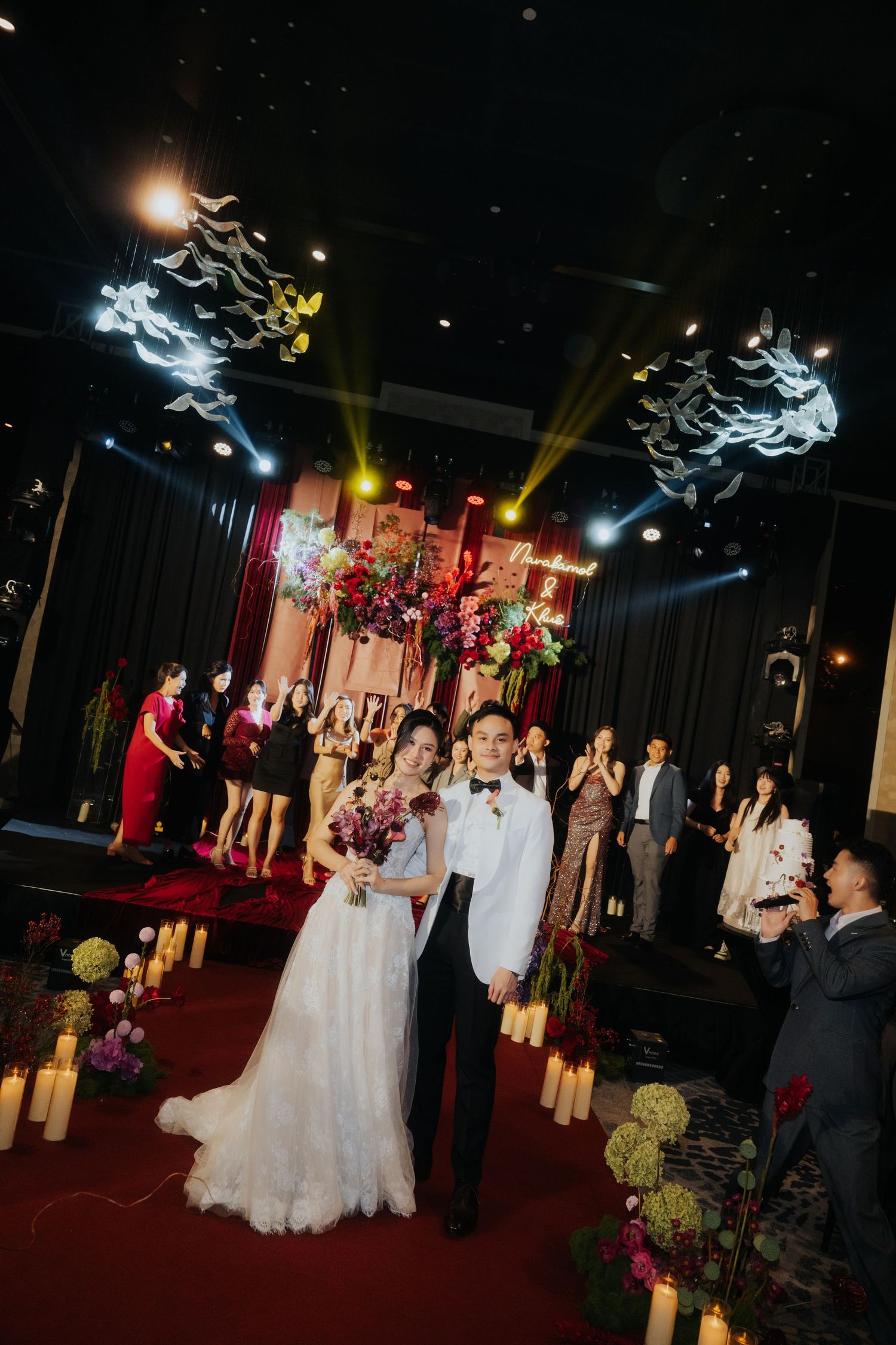 Bride and groom walk down candlelit aisle at elegant indoor wedding ceremony with floral decor and dramatic stage lighting.