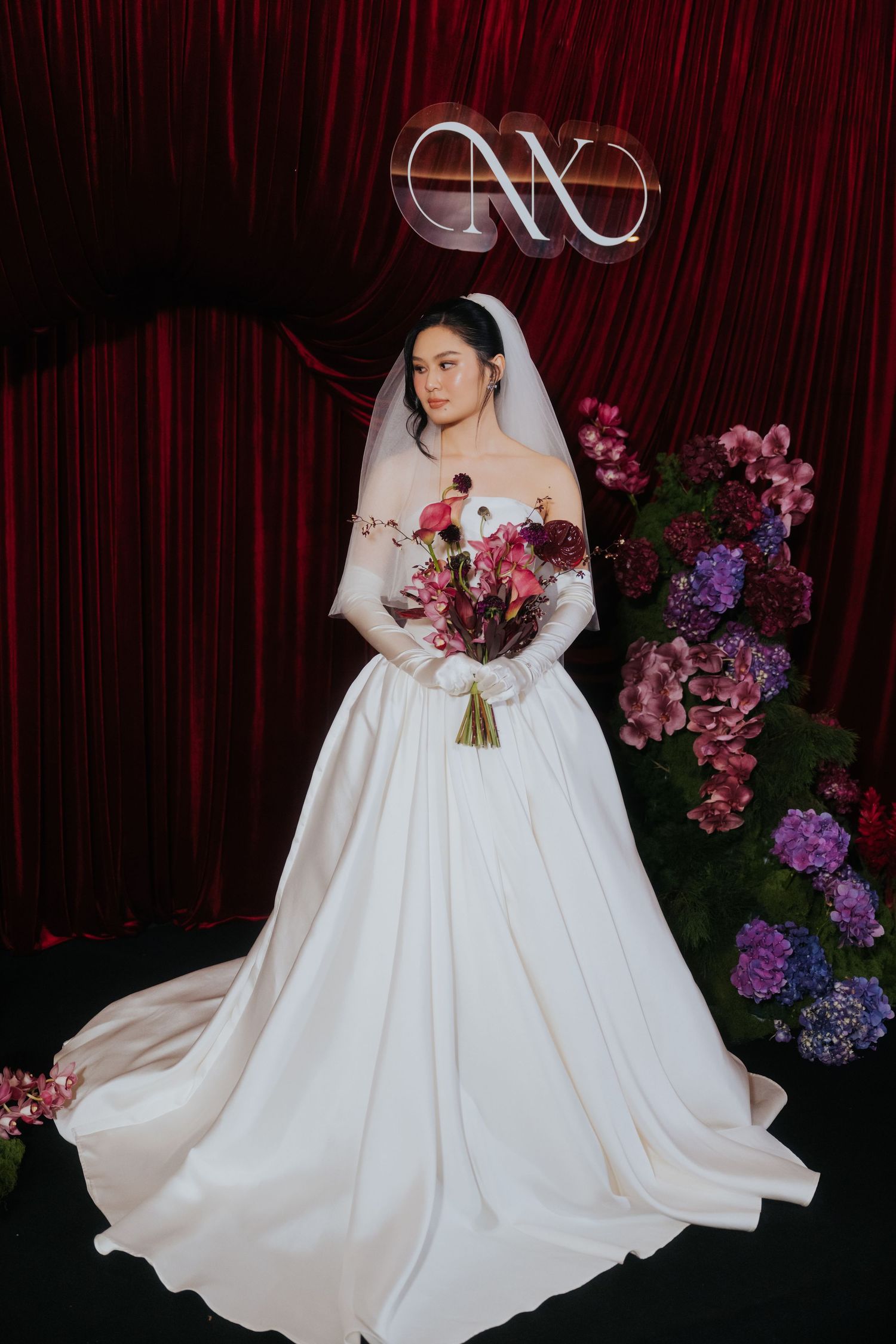 Bride in elegant white ball gown with veil holds bouquet of red and purple flowers before red curtain backdrop.