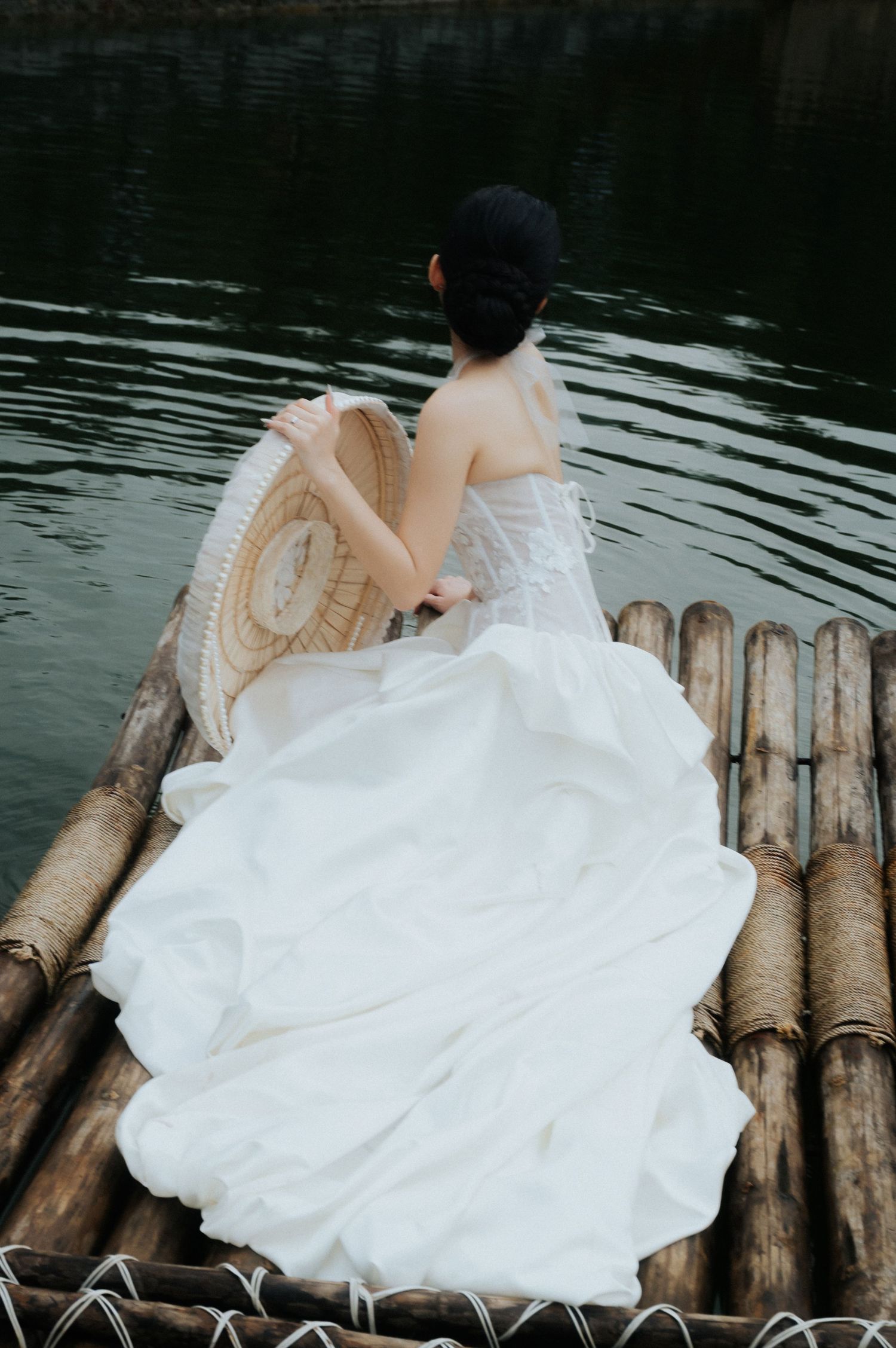 Woman in white wedding dress sitting on bamboo raft holding woven fan, facing calm dark water.