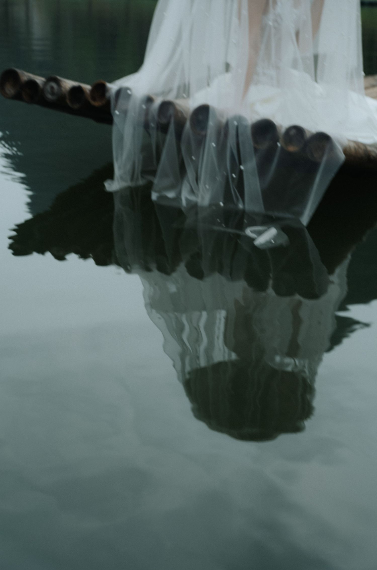 A bride in a white gown sits on a dock, her veil and dress reflecting beautifully in the calm, dark water below.