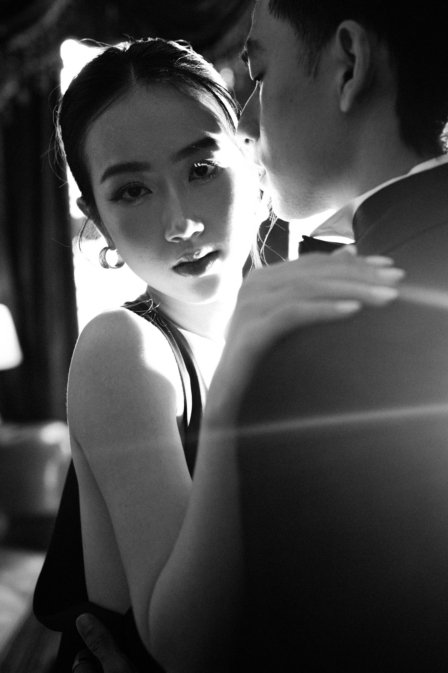 Black and white romantic portrait of a young Asian woman in a black dress gazing up at a man in soft dramatic lighting.