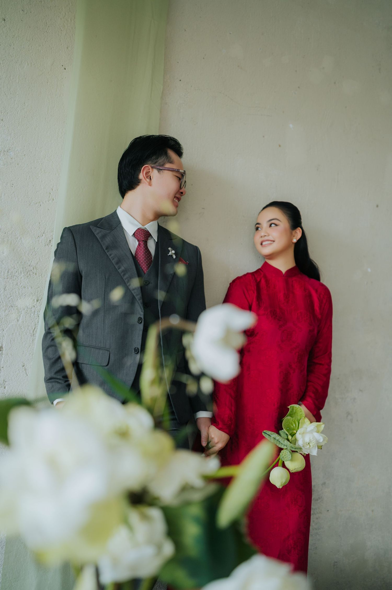 A couple in formal attire, man in gray suit and woman in red ao dai dress, holding hands and smiling at each other.