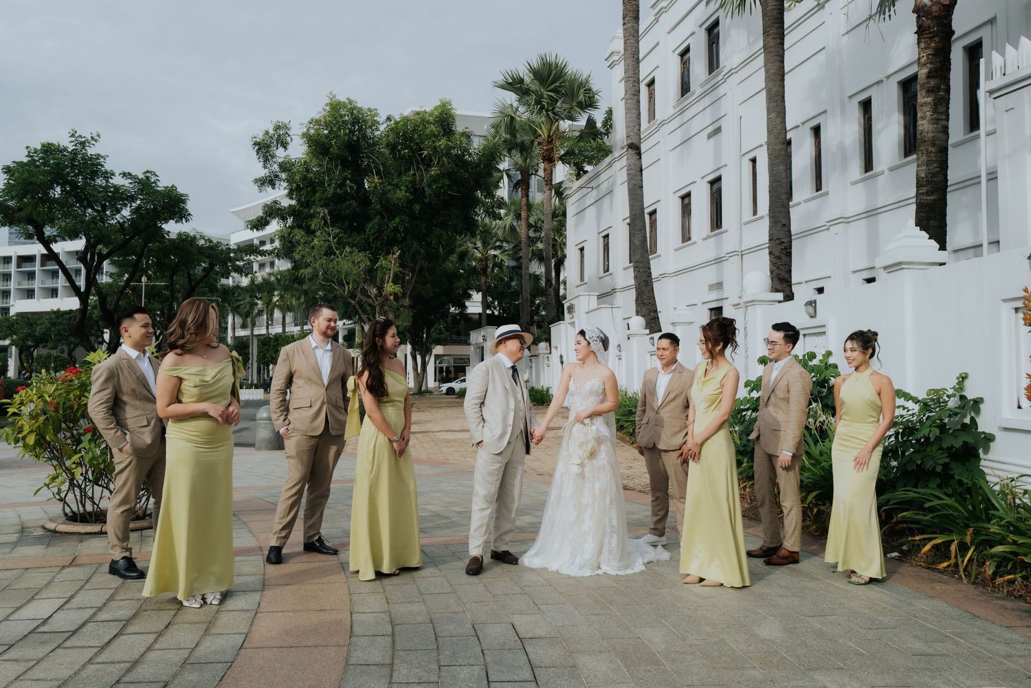 Wedding party in yellow bridesmaid dresses and tan suits stand outside a white building on a sunny day.