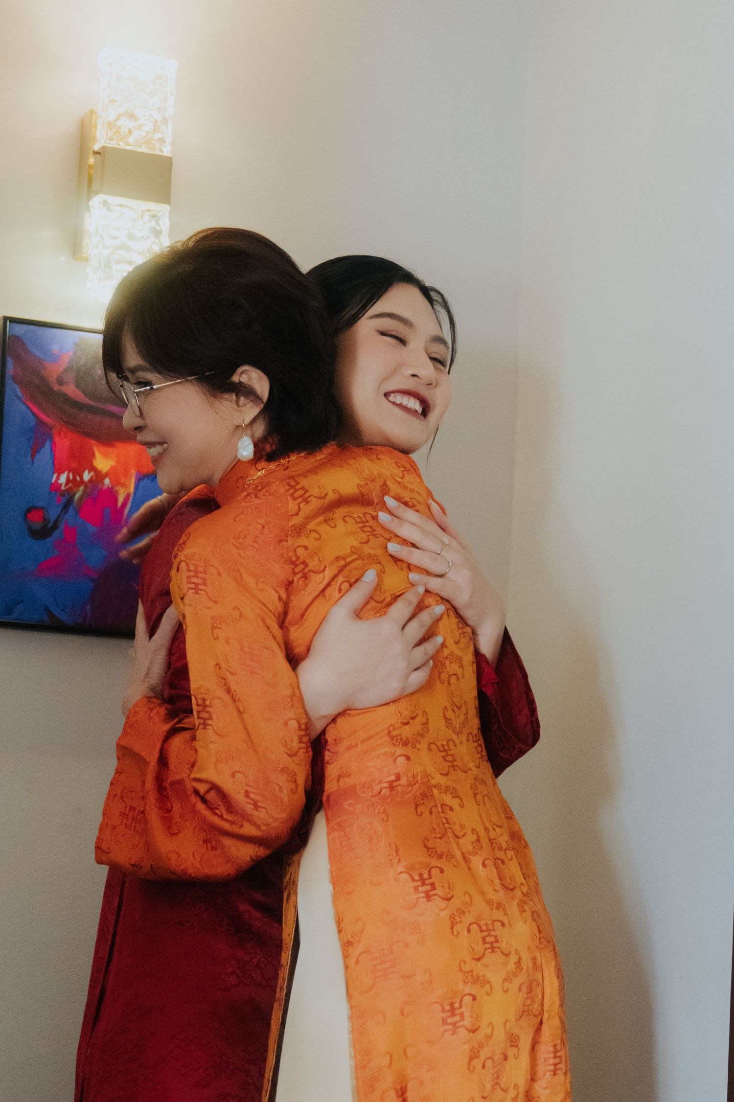 Two women in traditional orange and red Chinese qipao dresses share a warm embrace, smiling joyfully indoors.