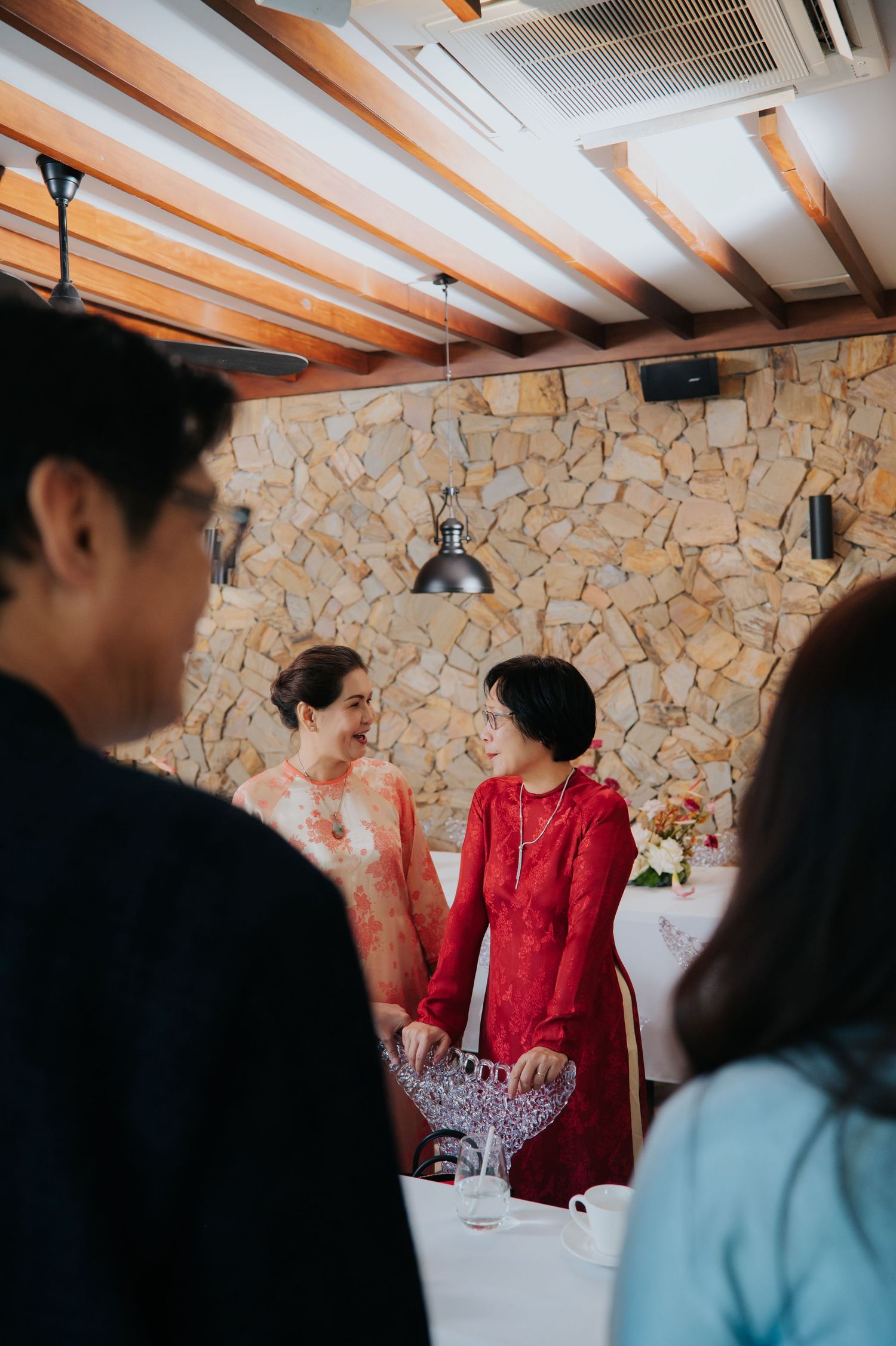 Two women in red traditional ao dai dresses stand together at a formal event in a stone-walled venue.