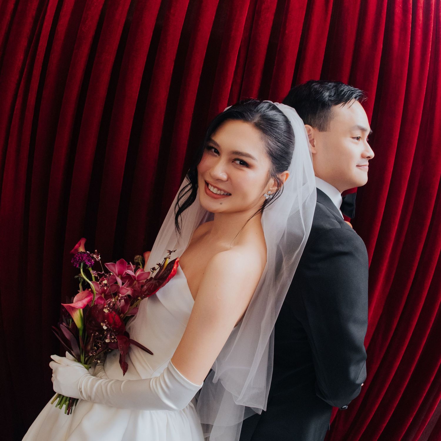 Bride in white gown and veil holds red bouquet, standing back-to-back with groom in suit before red curtain.