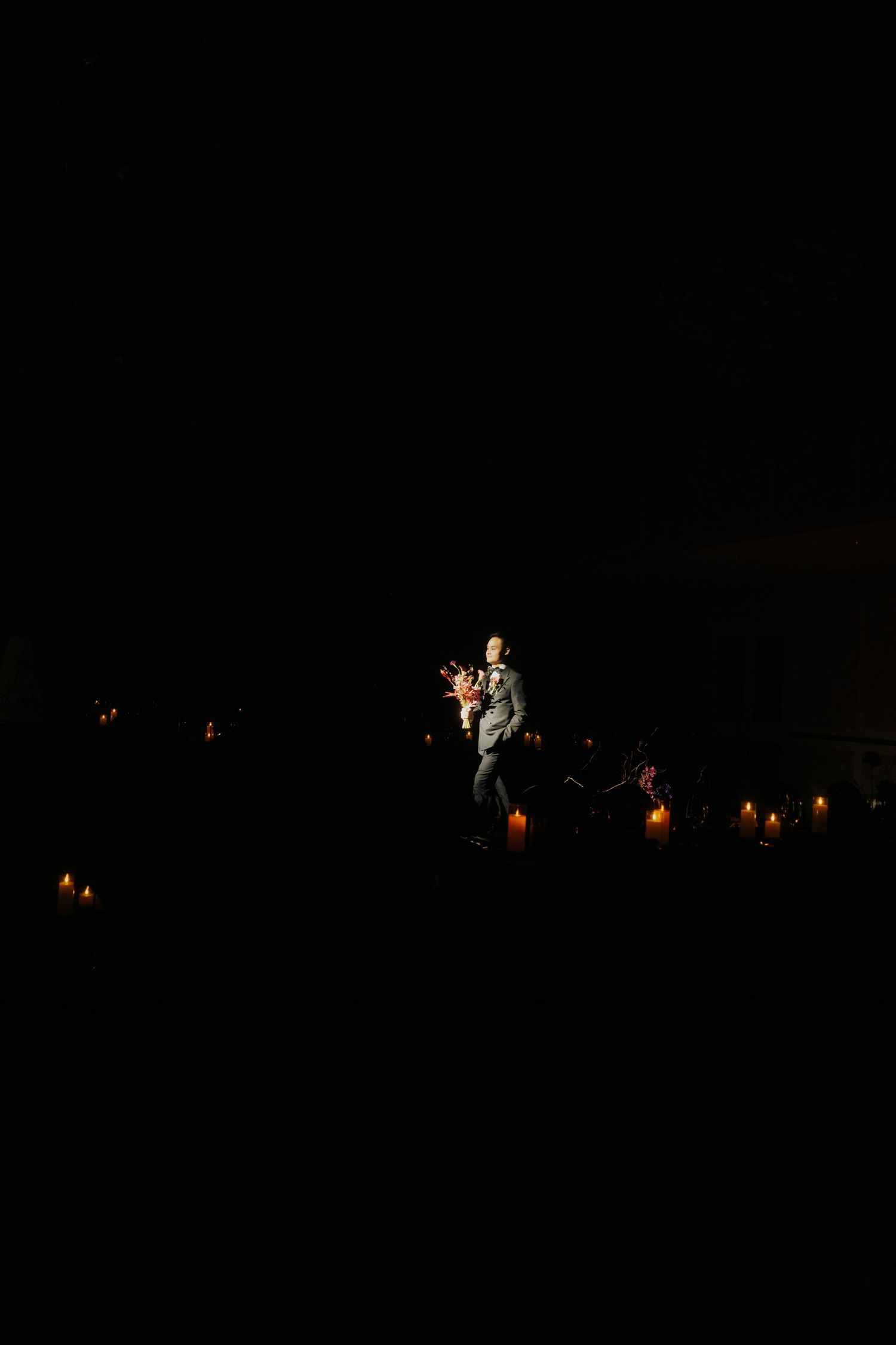 A lone performer stands in a spotlight on a dark stage surrounded by flickering candles during an intimate concert.