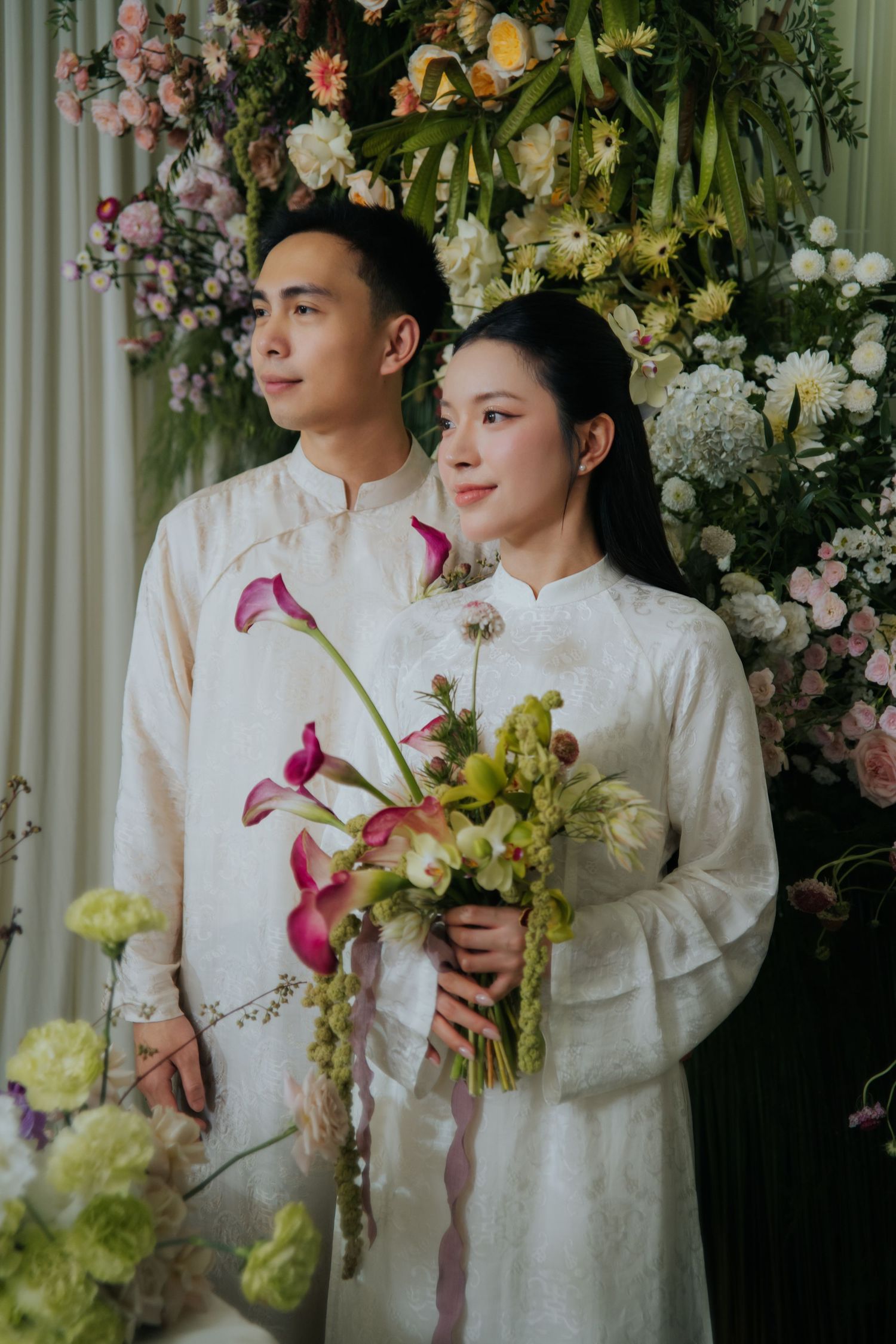 Vietnamese couple in white ao dai traditional attire pose with pink calla lily bouquet before floral backdrop.