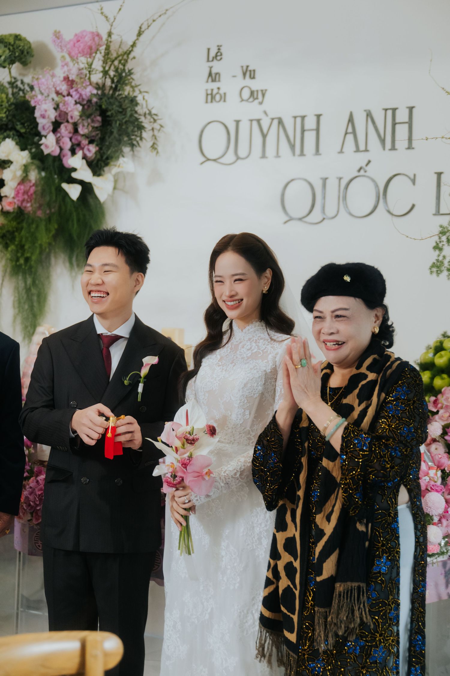 Vietnamese wedding ceremony with bride in white lace gown, groom in black suit, and elderly guest in traditional attire.