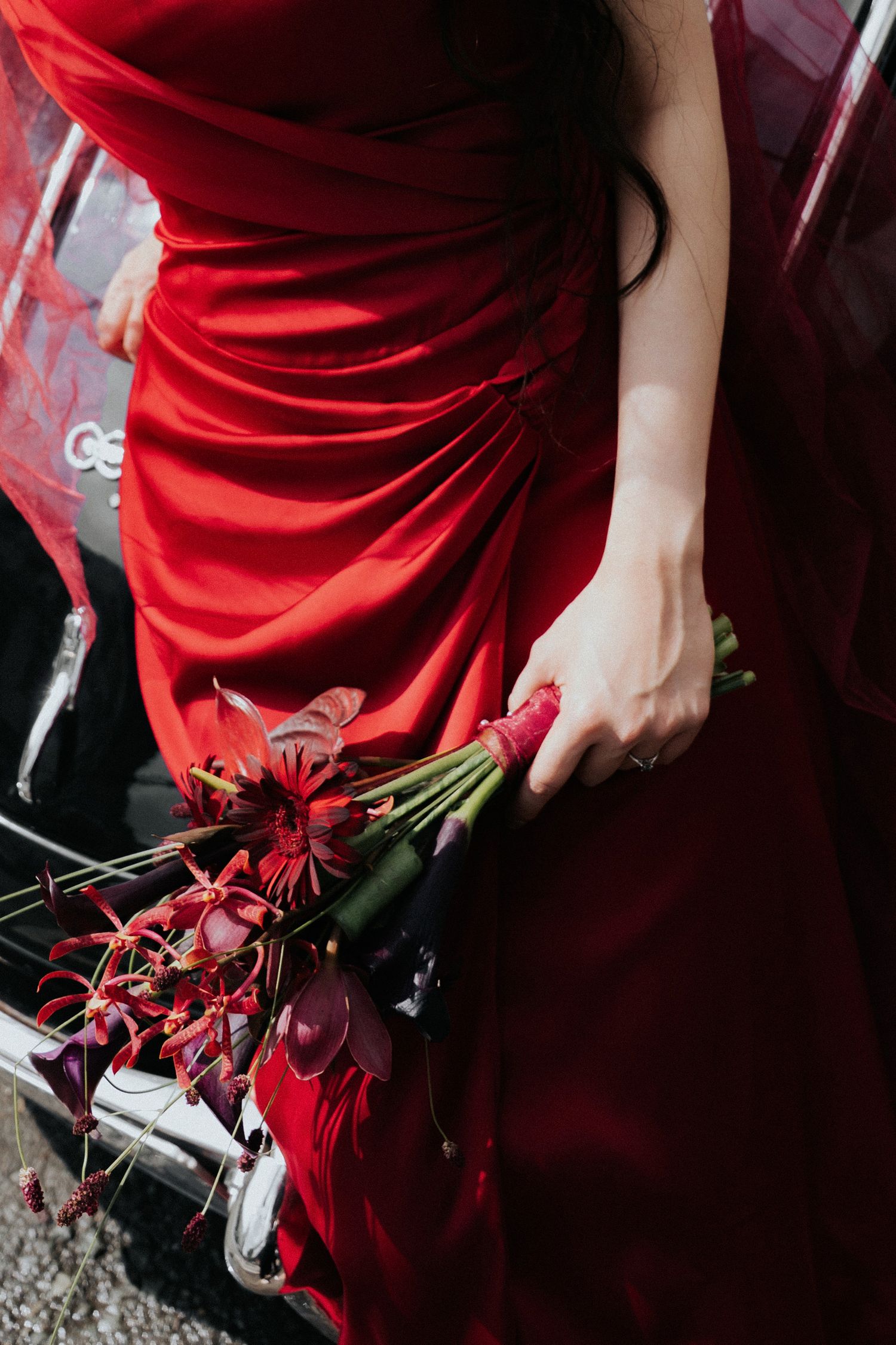 Woman in red ruched gown holding deep red floral bouquet beside a red car.