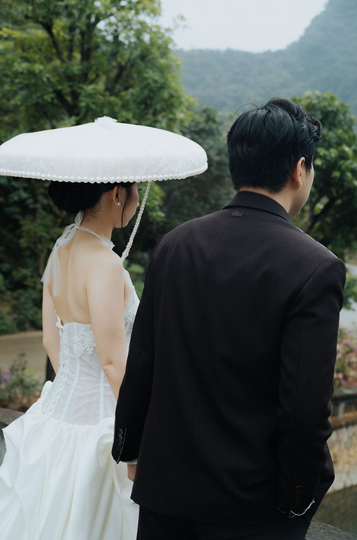 A bride in a white gown holding a lace umbrella stands beside a groom in a black suit, both facing a lush green landscape.