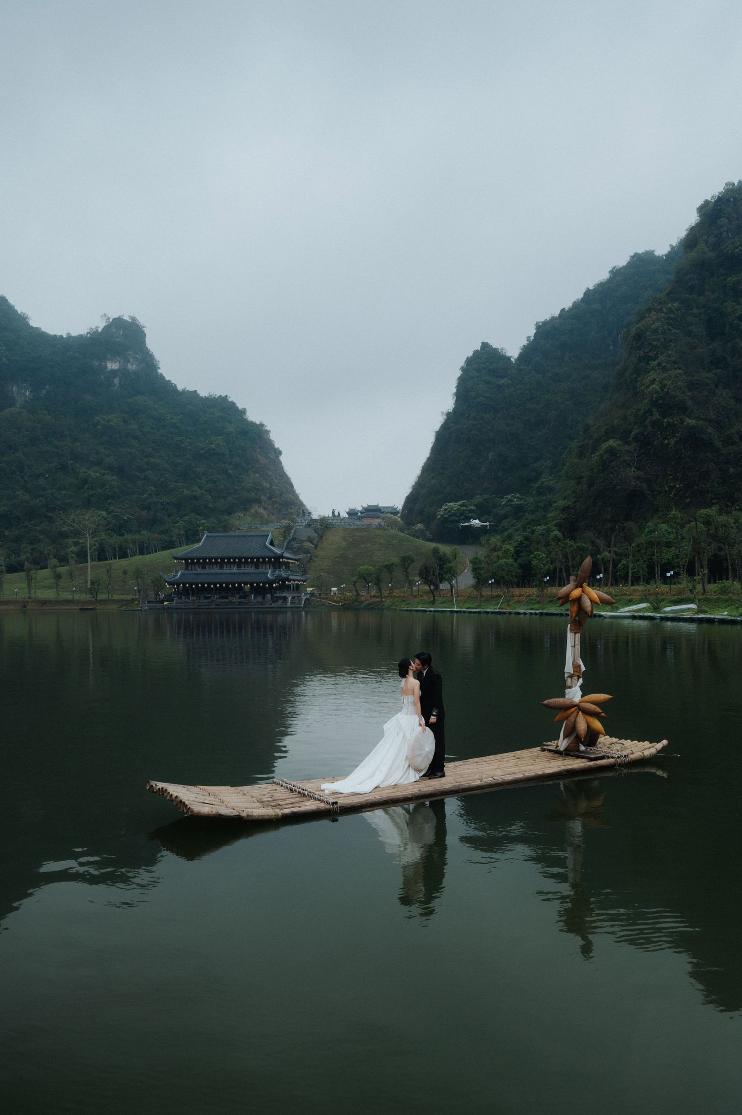 A couple in wedding attire sits on a bamboo raft on a serene lake surrounded by karst mountains in Vietnam.