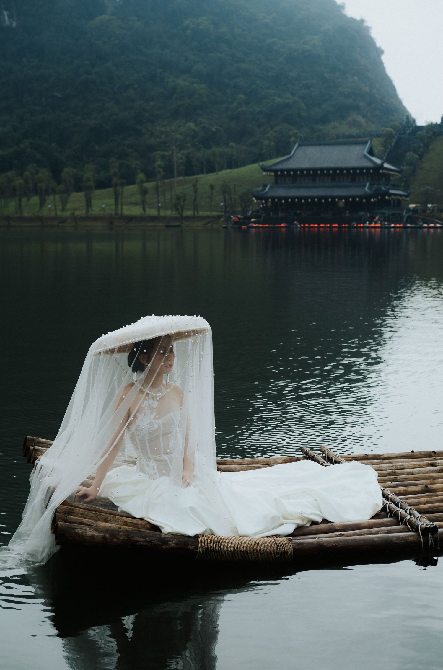 Bride in white gown and veil sits on bamboo raft on calm lake with pagoda and green hills behind.