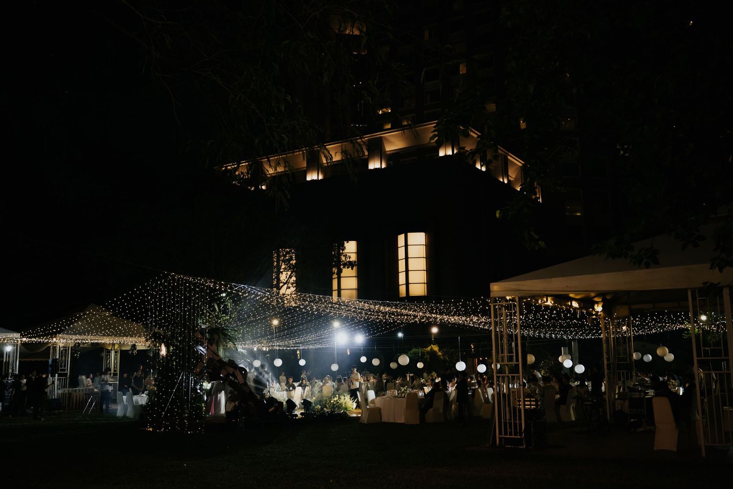 Elegant outdoor nighttime event with string lights and white decorations illuminating a garden venue beneath a lit building.