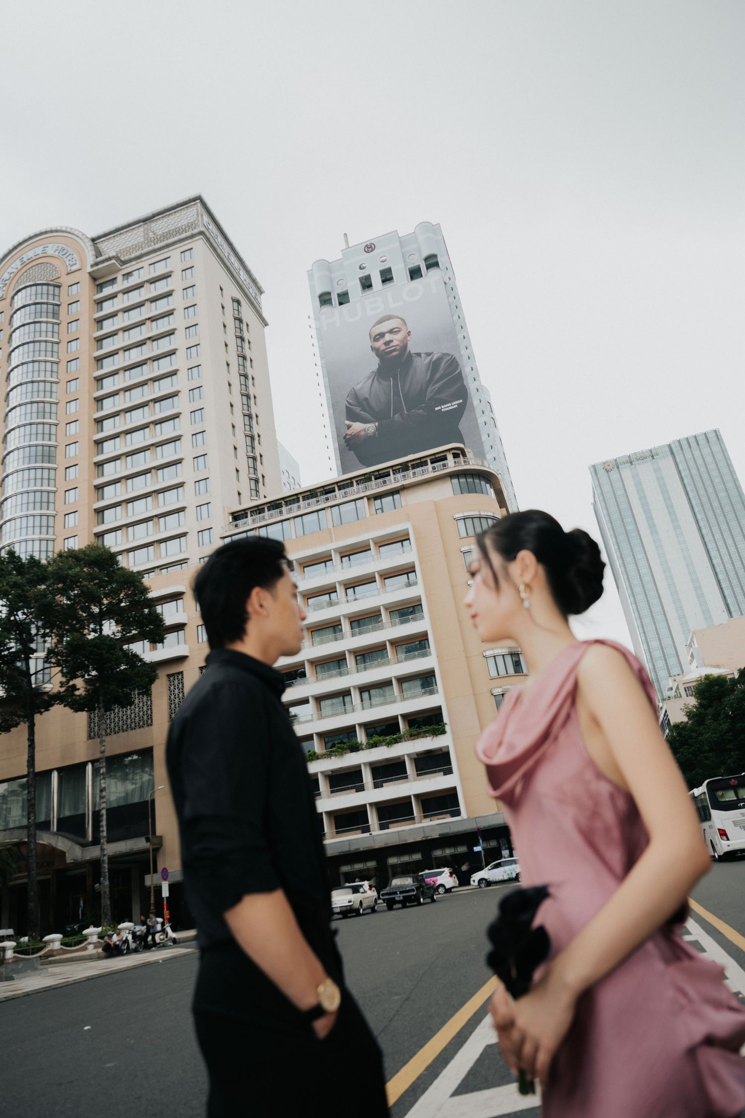 Couple in formal attire stand on a city street with a large billboard and tall buildings in the background.