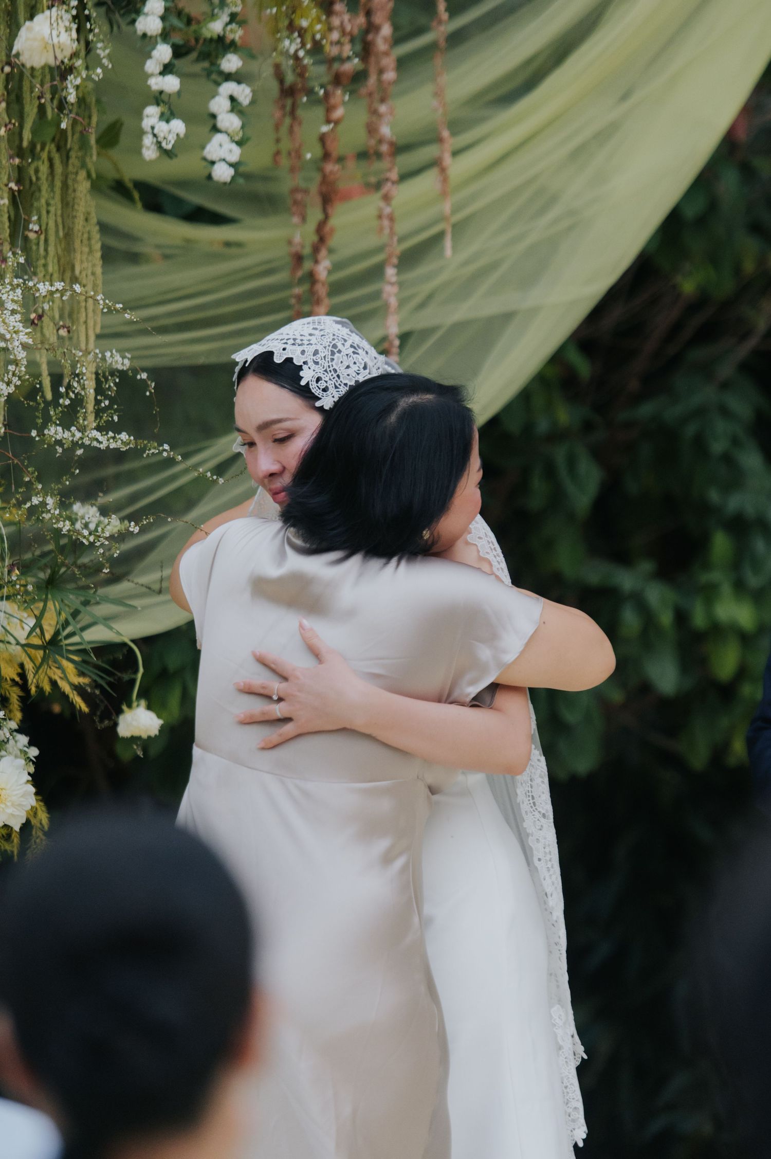 A bride in a white gown and lace headpiece shares an emotional hug at an outdoor wedding ceremony.