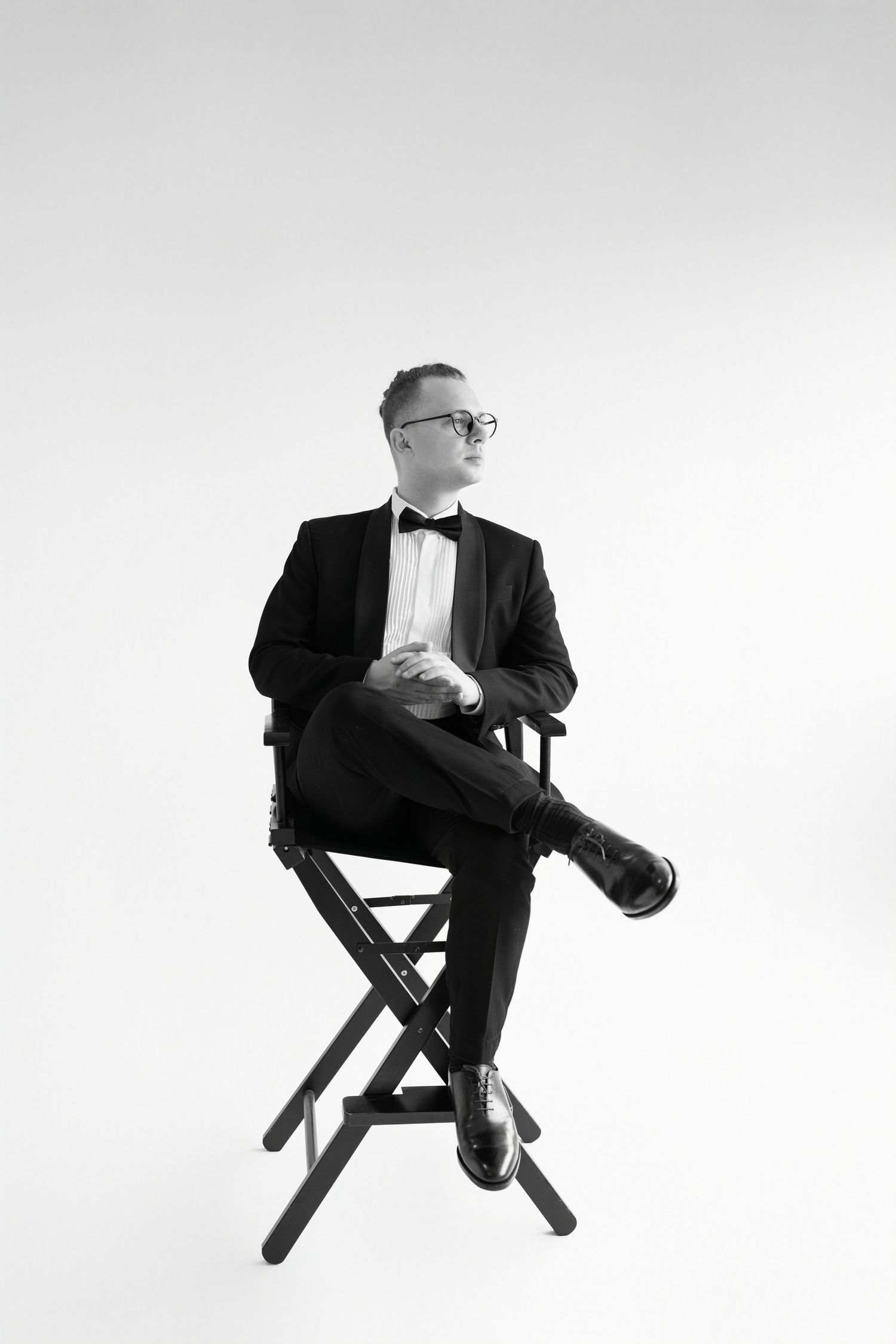 Black and white photo of a man in a tuxedo and bow tie sitting in a director's chair with legs crossed.