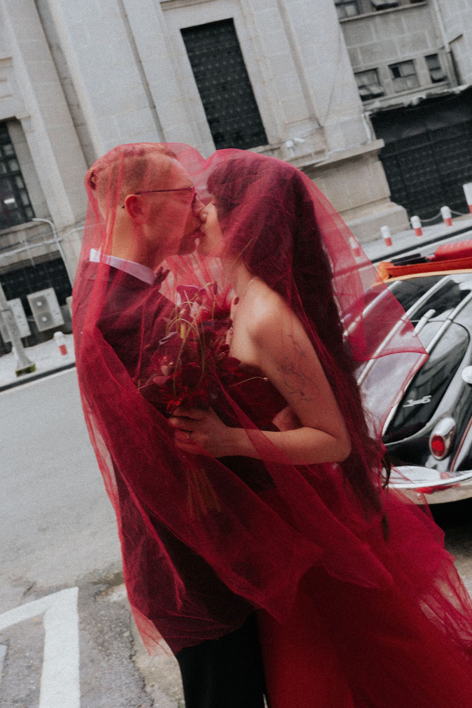 Couple kissing under a dramatic red tulle veil, bride holding bouquet, urban street backdrop.