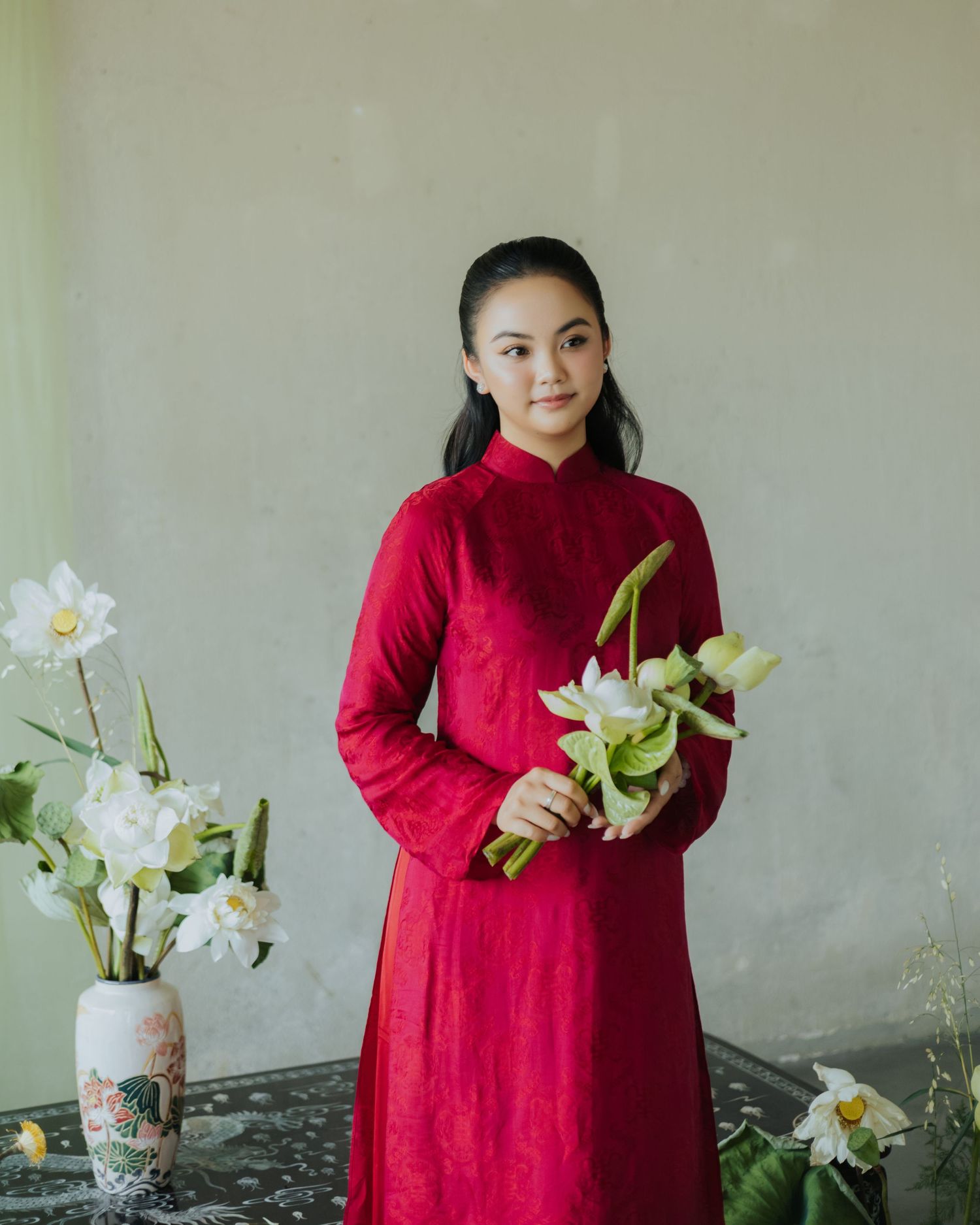 Young Vietnamese woman in red ao dai holding white lotus flowers, standing beside a floral vase against a soft green wall.