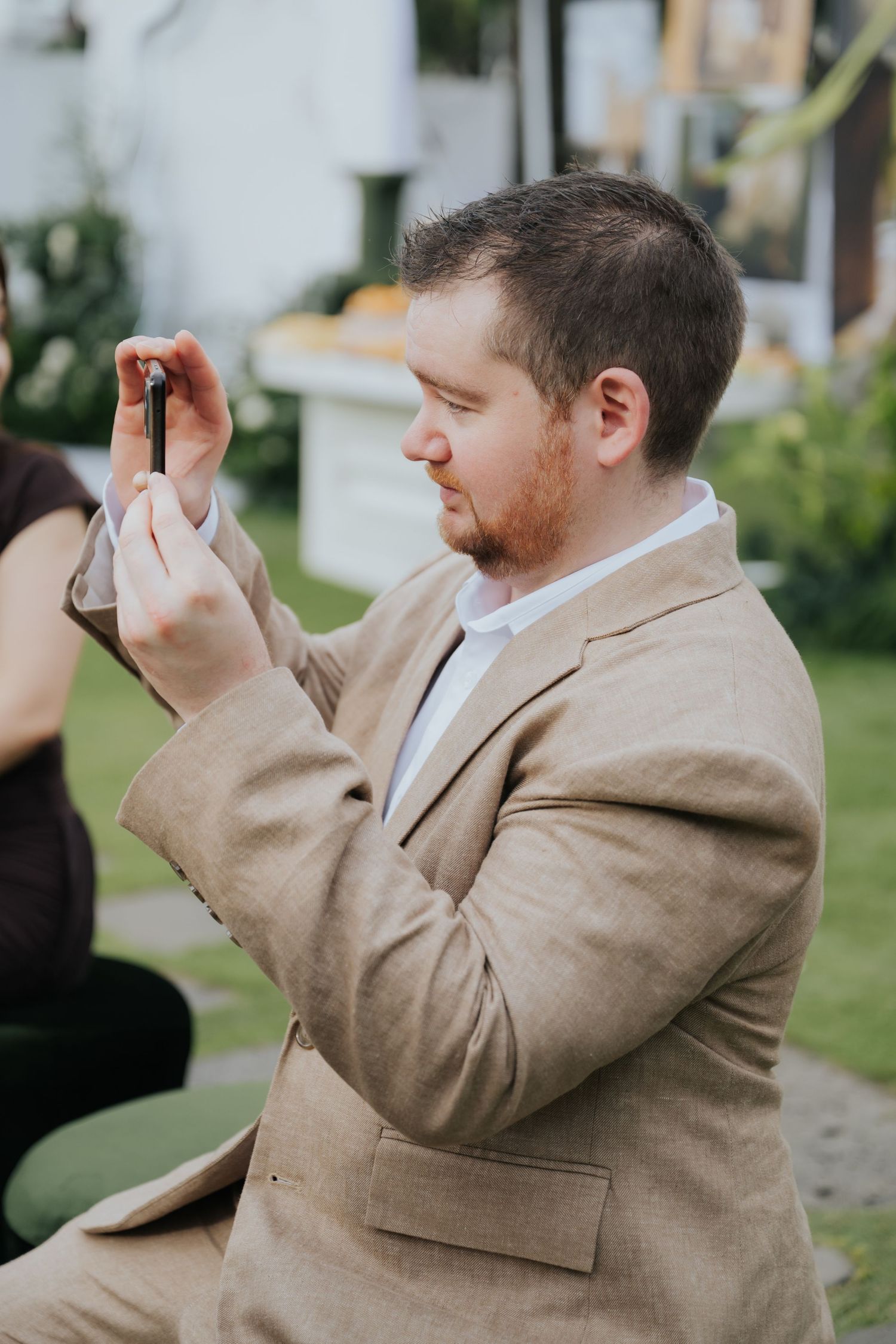 Man in beige suit jacket taking a photo with his smartphone outdoors at a garden event.