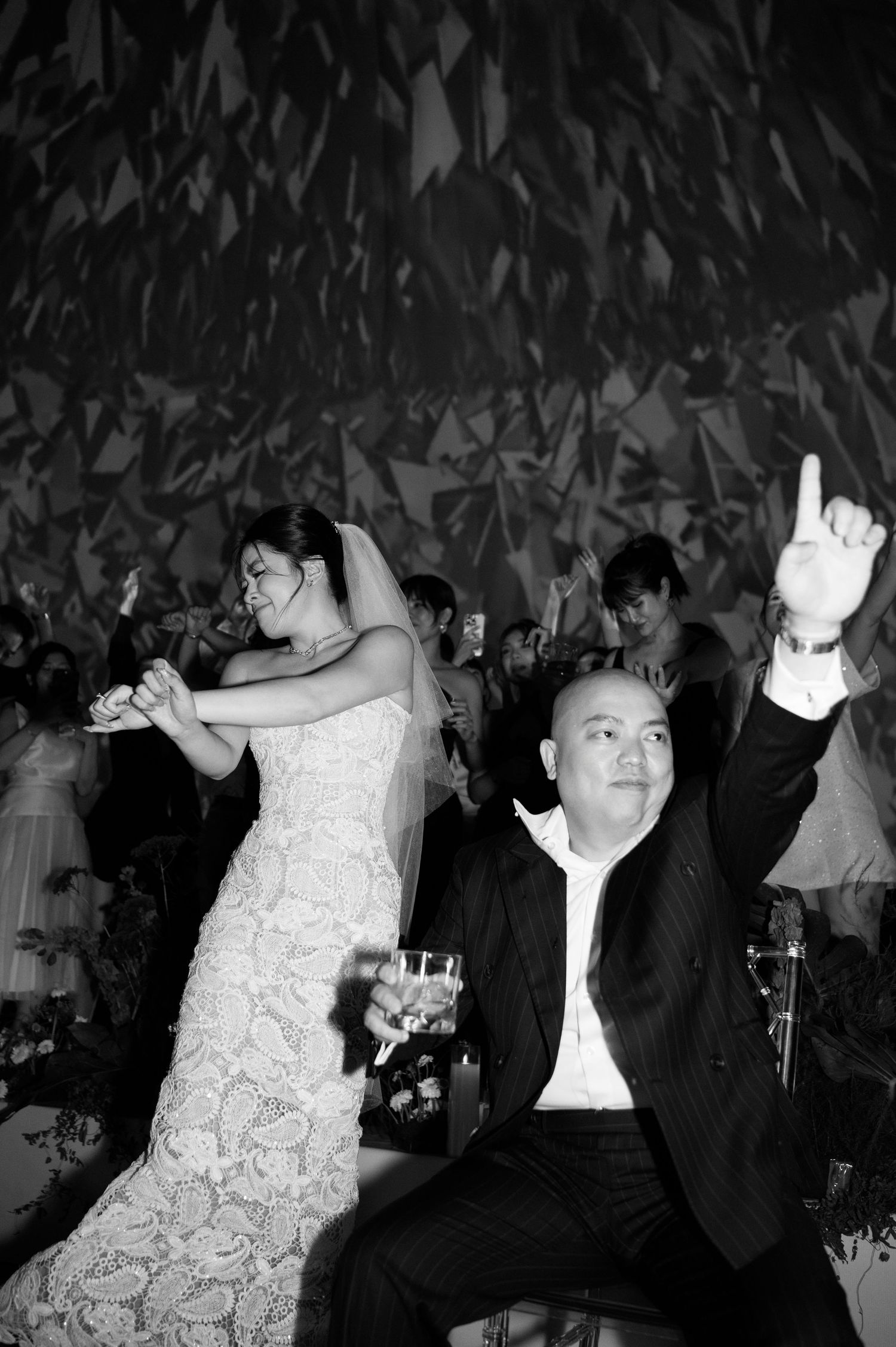 Black and white photo of a bride in lace gown and groom in suit celebrating joyfully at their wedding reception.