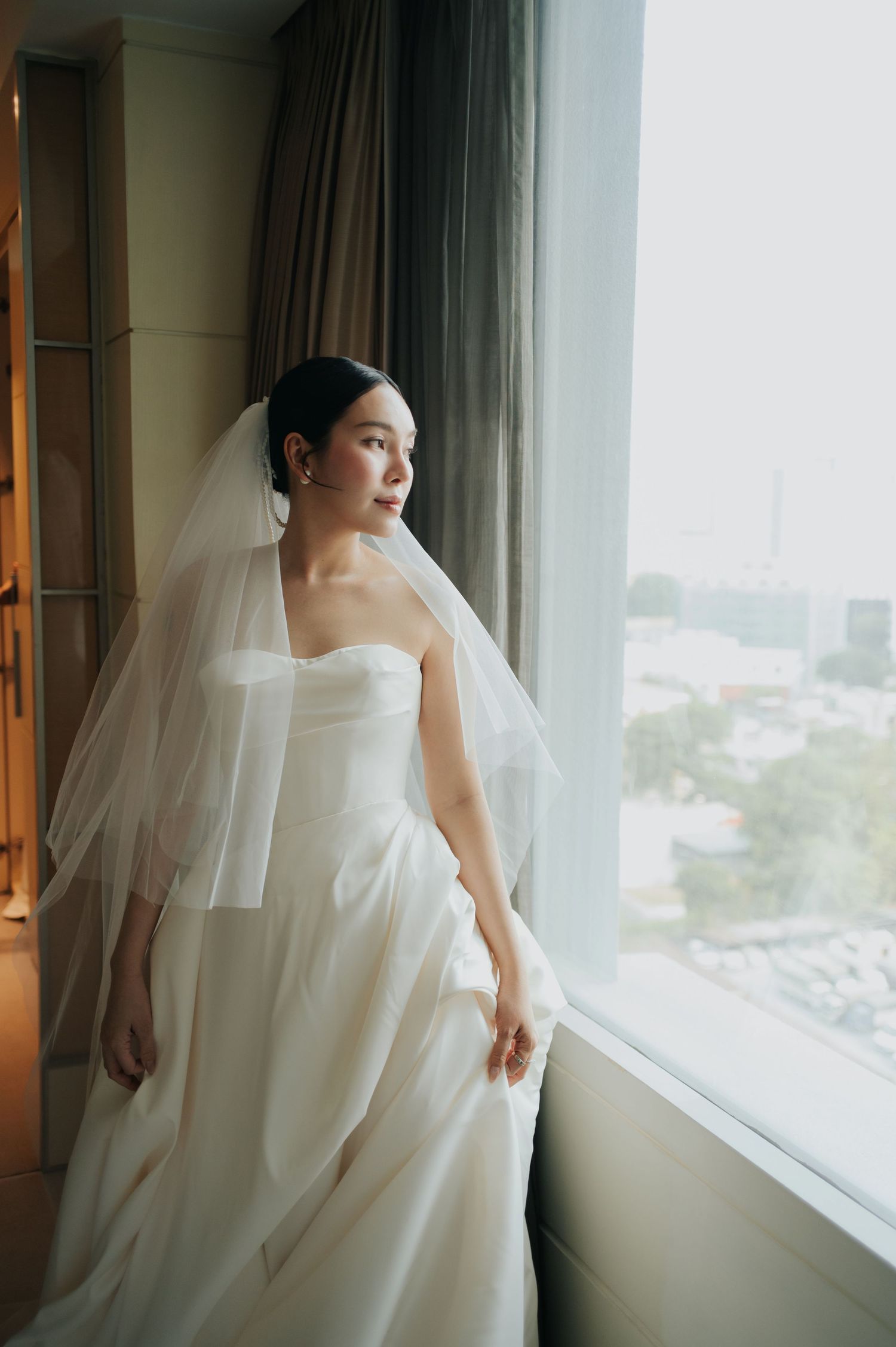 A bride in a strapless white gown and veil gazes out a large window, bathed in soft natural light.