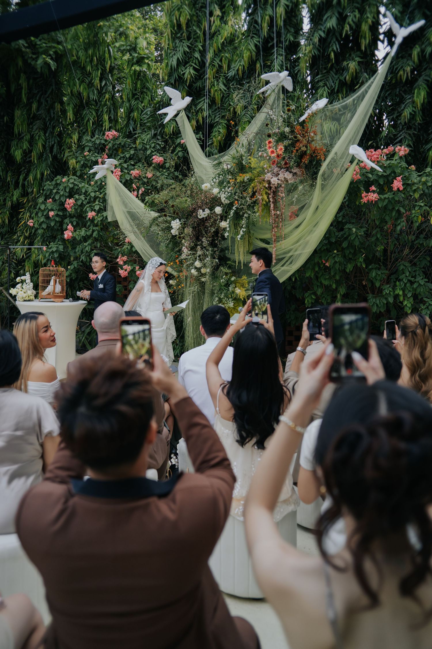 Outdoor wedding ceremony with guests photographing couple at floral arch with green draping and white doves.