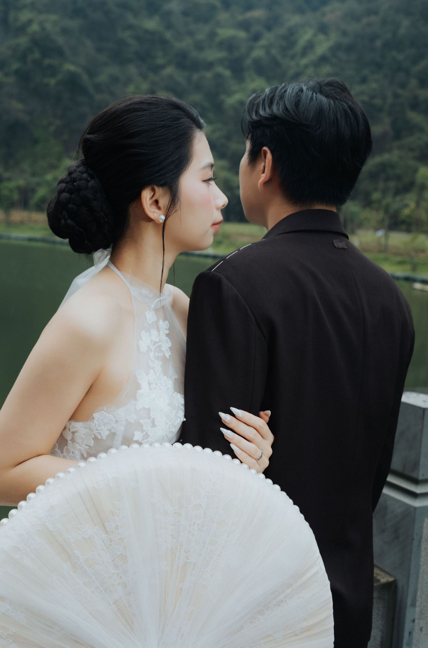 A bride in white lace dress and groom in dark suit share an intimate moment outdoors near a lake.