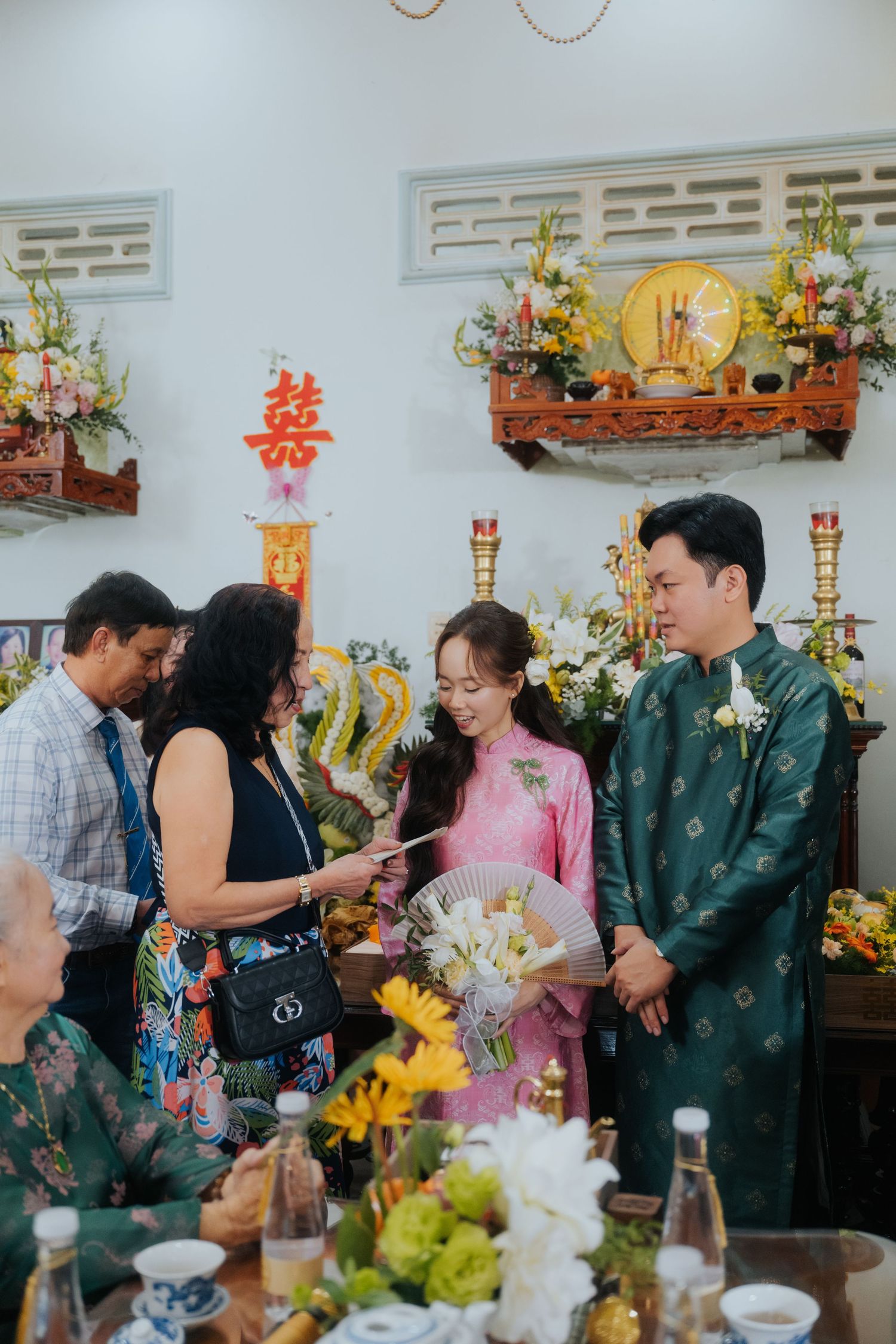 Vietnamese couple in traditional ao dai attire during a wedding ceremony with family and floral decorations.