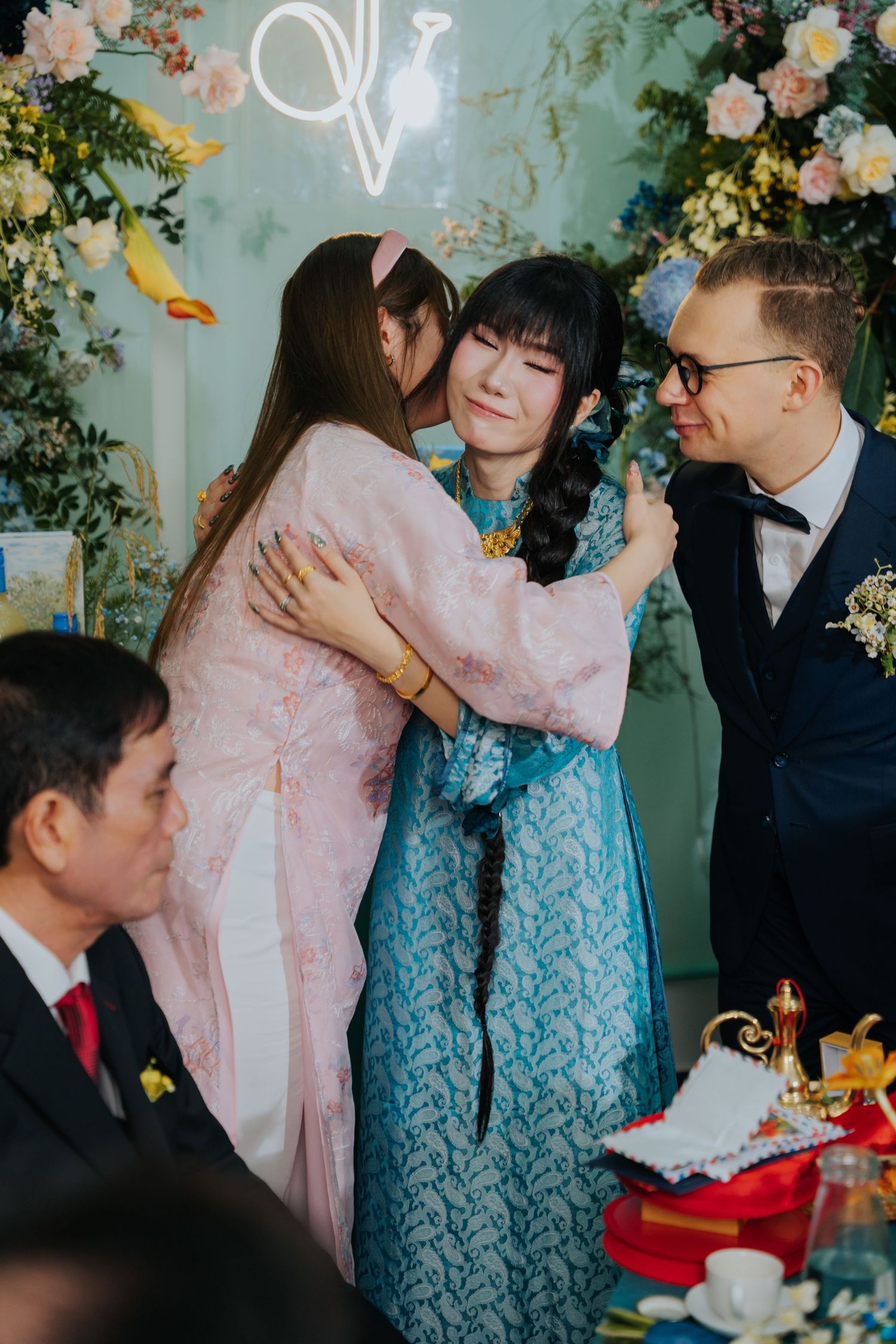 Two women embrace warmly at a wedding reception, with a groom in a navy suit smiling nearby amid floral decor.