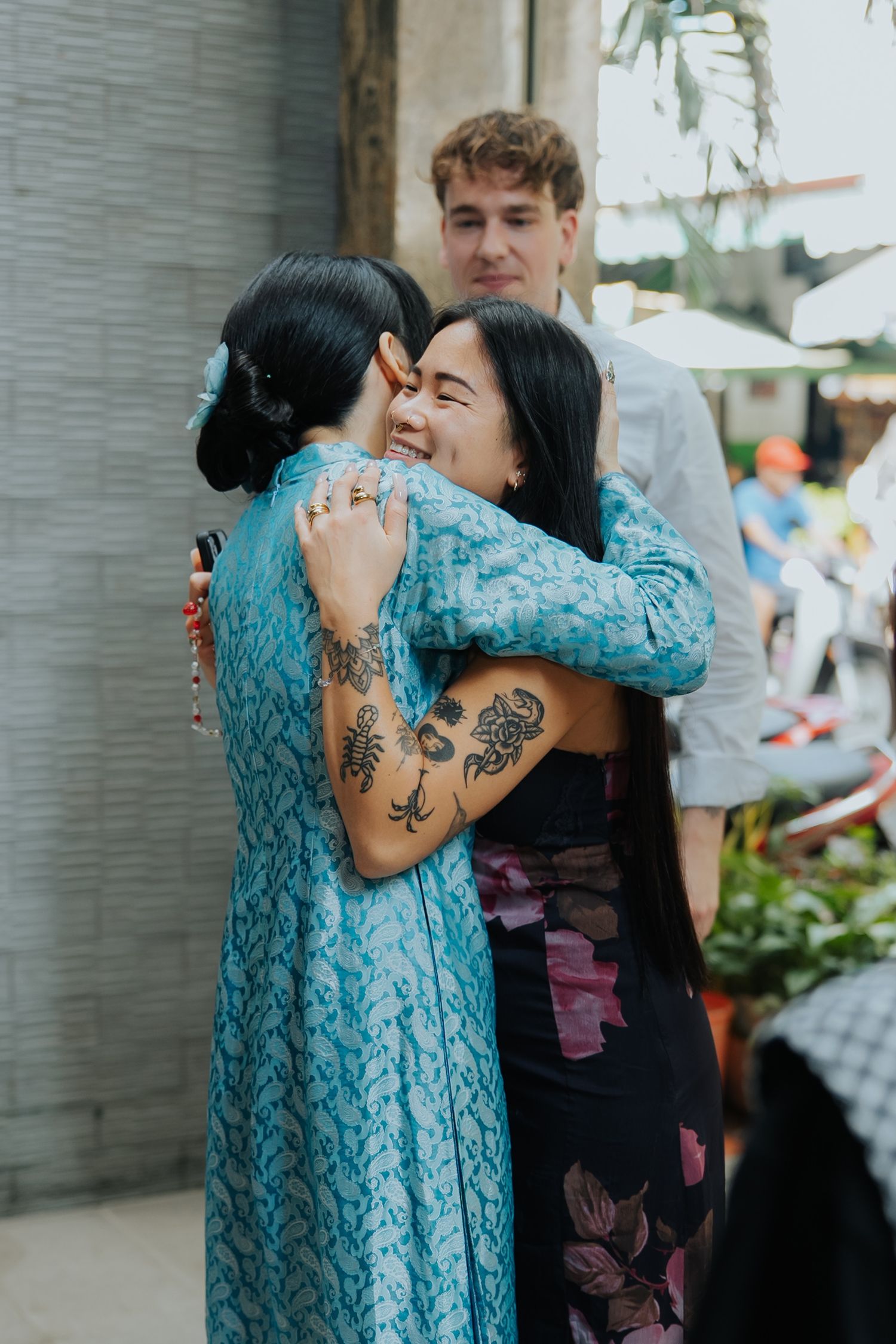 Two women with tattoos share a warm hug outdoors, wearing blue patterned dress, with a man standing behind them.